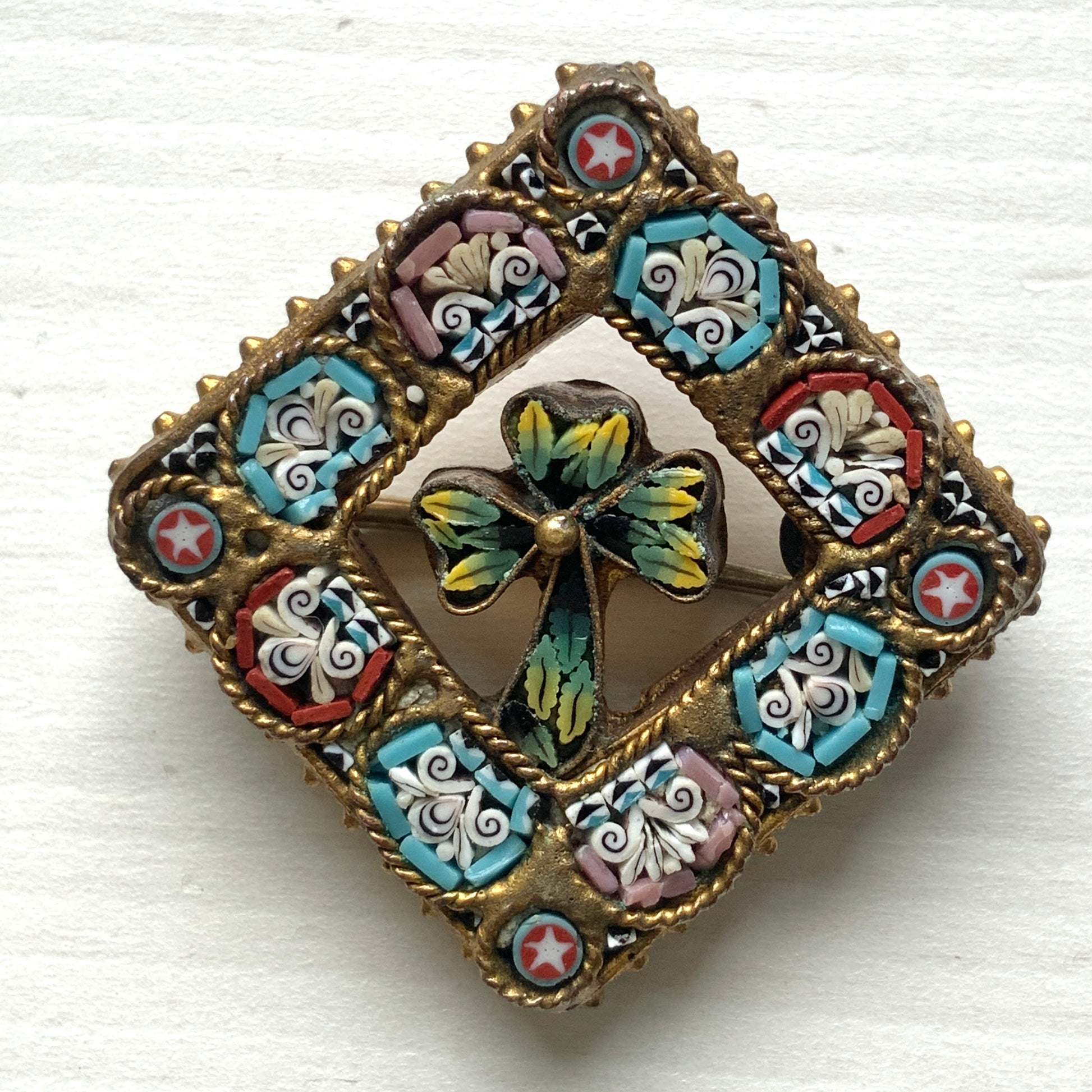 A brooch featuring a three-leaf clover set inside a diamond-shaped frame adorned with delicate detailing. 細かな飾りが飾られたダイヤ型のフレームの中に、三つ葉のクローバーが入ったブローチ。