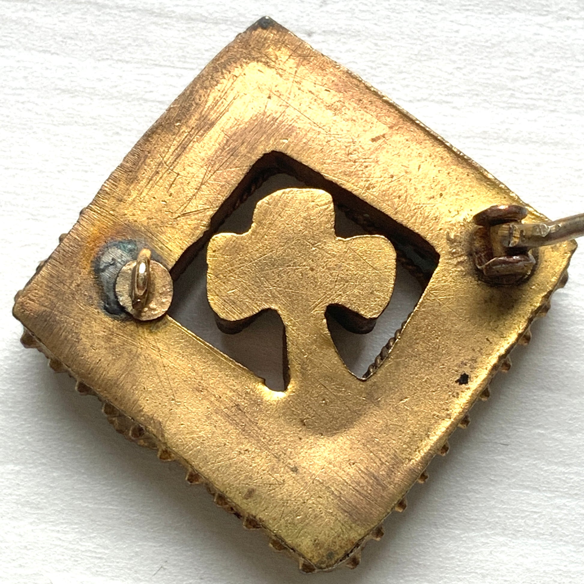 A brooch featuring a three-leaf clover set inside a diamond-shaped frame adorned with delicate detailing. 細かな飾りが飾られたダイヤ型のフレームの中に、三つ葉のクローバーが入ったブローチ。