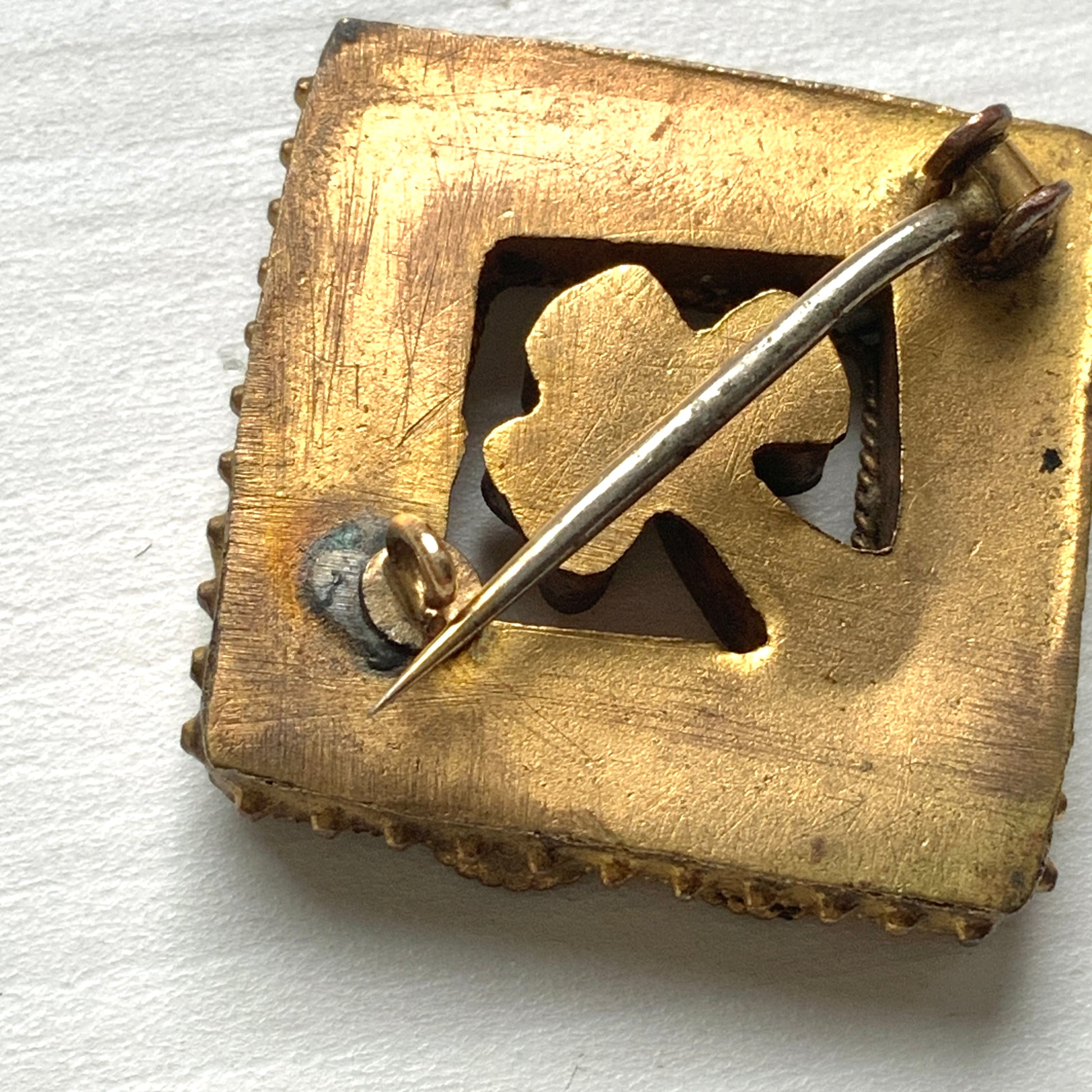 A brooch featuring a three-leaf clover set inside a diamond-shaped frame adorned with delicate detailing. 細かな飾りが飾られたダイヤ型のフレームの中に、三つ葉のクローバーが入ったブローチ。