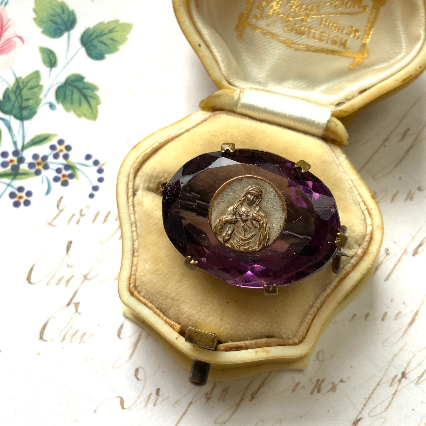 An antique brooch featuring a medal of the Virgin Mary set at the centre of a deep purple faceted glass stone. 深いパープルのカットガラスの中央に、聖母マリアを象ったメダルをセットしたアンティークブローチ。