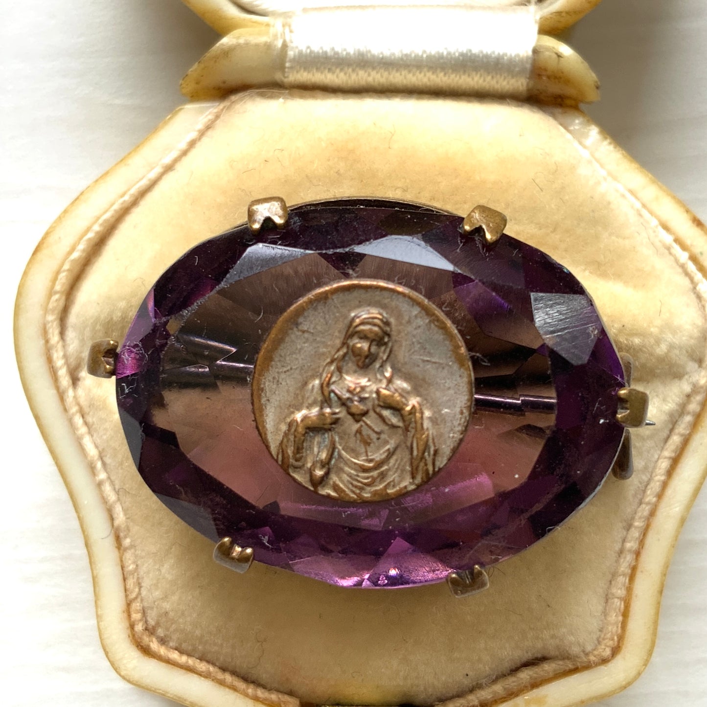 An antique brooch featuring a medal of the Virgin Mary set at the centre of a deep purple faceted glass stone. 深いパープルのカットガラスの中央に、聖母マリアを象ったメダルをセットしたアンティークブローチ。