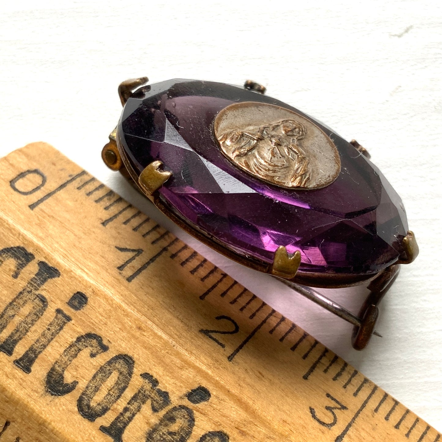 An antique brooch featuring a medal of the Virgin Mary set at the centre of a deep purple faceted glass stone. 深いパープルのカットガラスの中央に、聖母マリアを象ったメダルをセットしたアンティークブローチ。