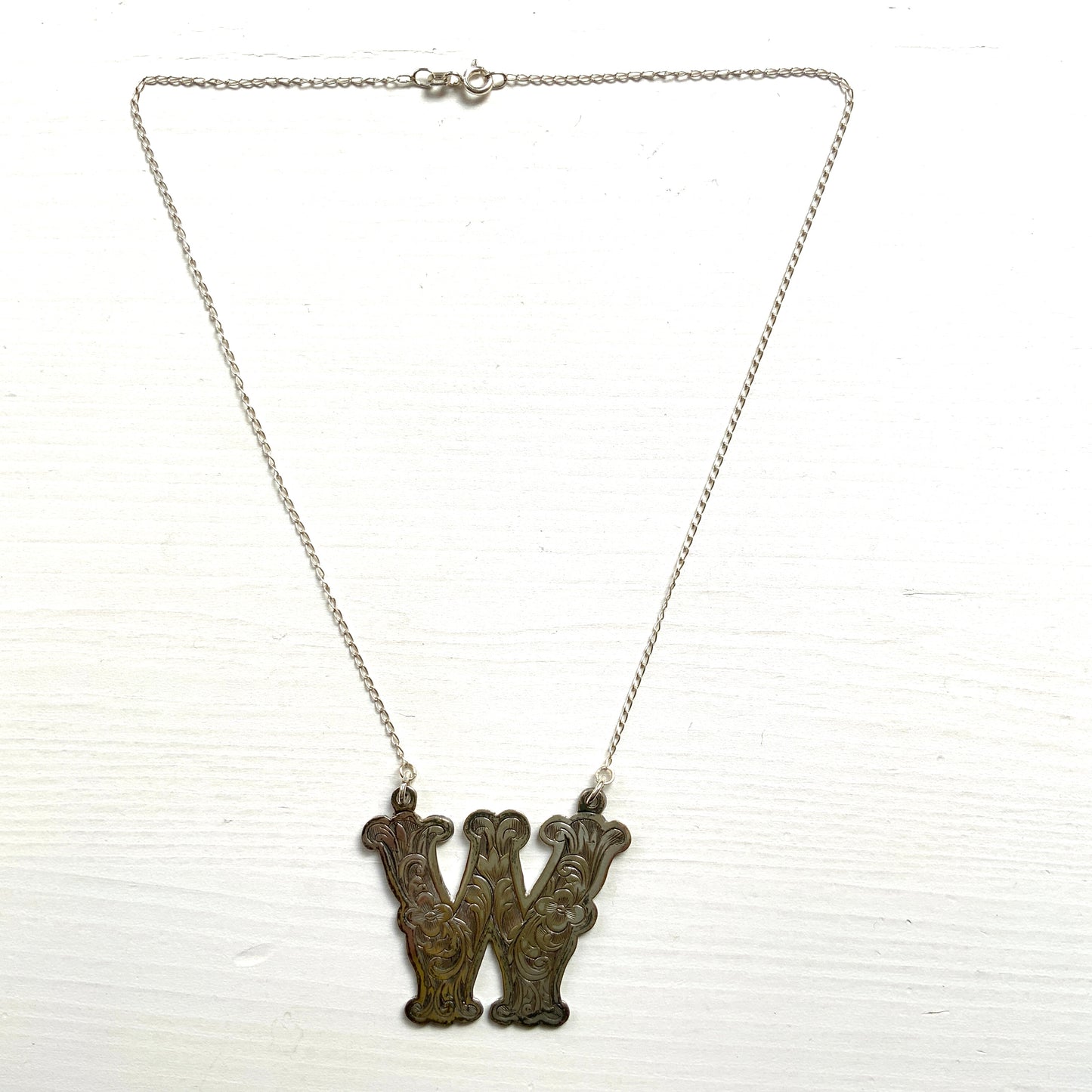 A necklace featuring the letter “W”, beautifully engraved with a botanical motif. 美しい植物モチーフのエングレービングが施された“W”のネックレス。