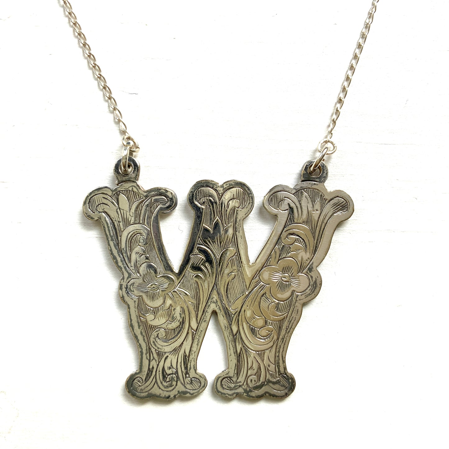 A necklace featuring the letter “W”, beautifully engraved with a botanical motif. 美しい植物モチーフのエングレービングが施された“W”のネックレス。