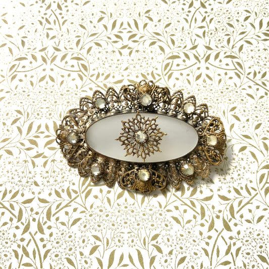 A brooch featuring a white oval frosted glass, framed with gold-tone filigree. 白いオーバルのフロスとグラスを、ゴールドトーンのフィリグリー縁取ったブローチ。