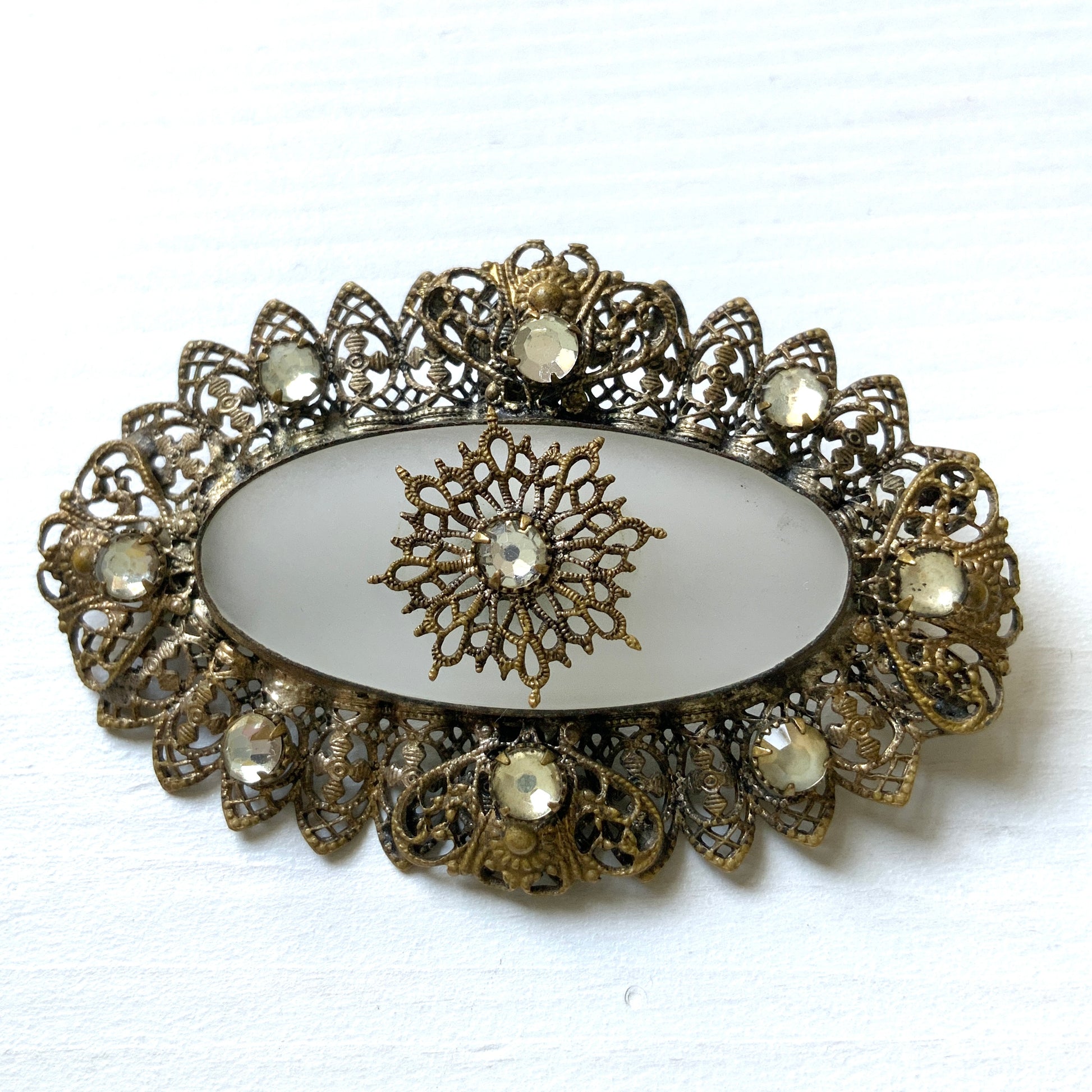 A brooch featuring a white oval frosted glass, framed with gold-tone filigree. 白いオーバルのフロスとグラスを、ゴールドトーンのフィリグリー縁取ったブローチ。