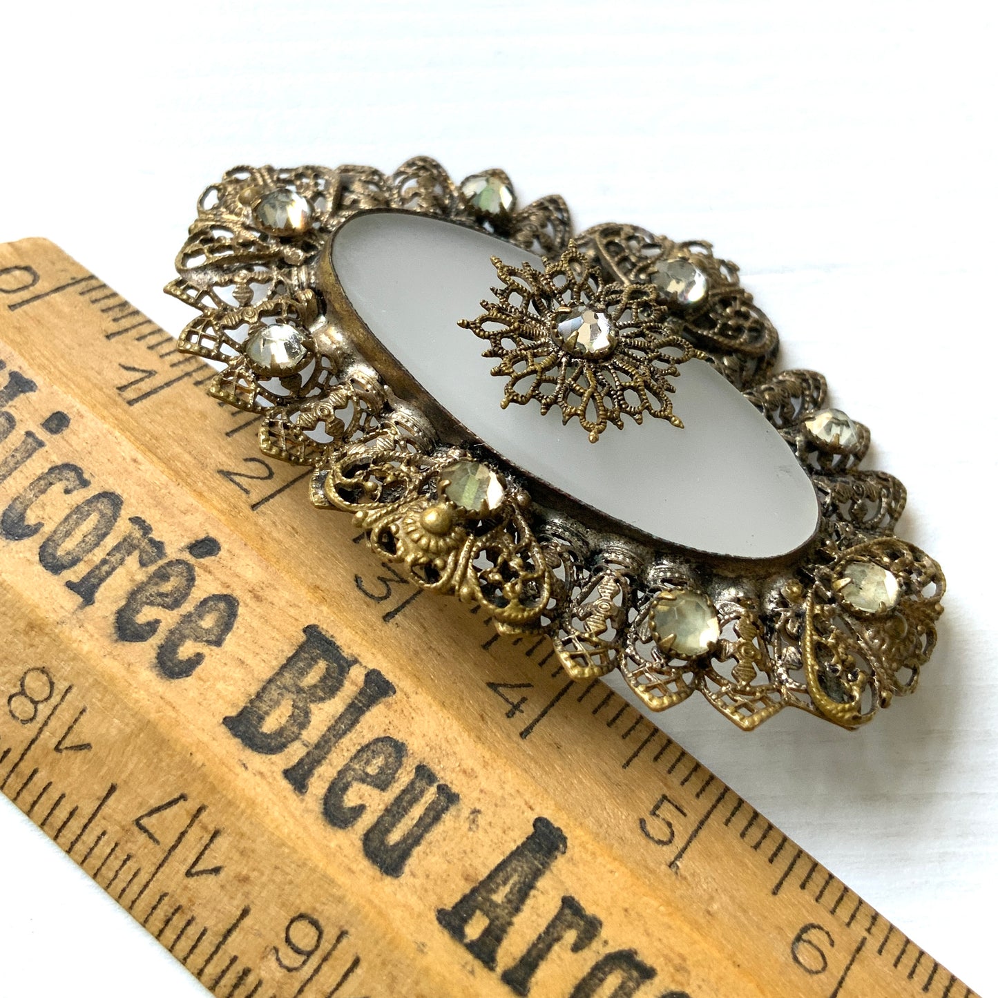A brooch featuring a white oval frosted glass, framed with gold-tone filigree. 白いオーバルのフロスとグラスを、ゴールドトーンのフィリグリー縁取ったブローチ。