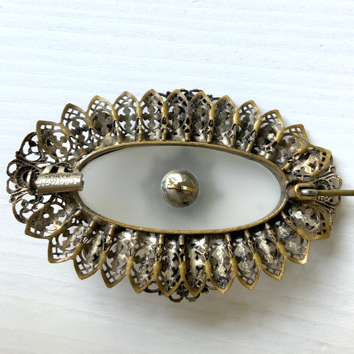 A brooch featuring a white oval frosted glass, framed with gold-tone filigree. 白いオーバルのフロスとグラスを、ゴールドトーンのフィリグリー縁取ったブローチ。