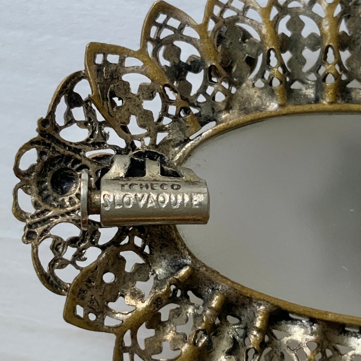 A brooch featuring a white oval frosted glass, framed with gold-tone filigree. 白いオーバルのフロスとグラスを、ゴールドトーンのフィリグリー縁取ったブローチ。