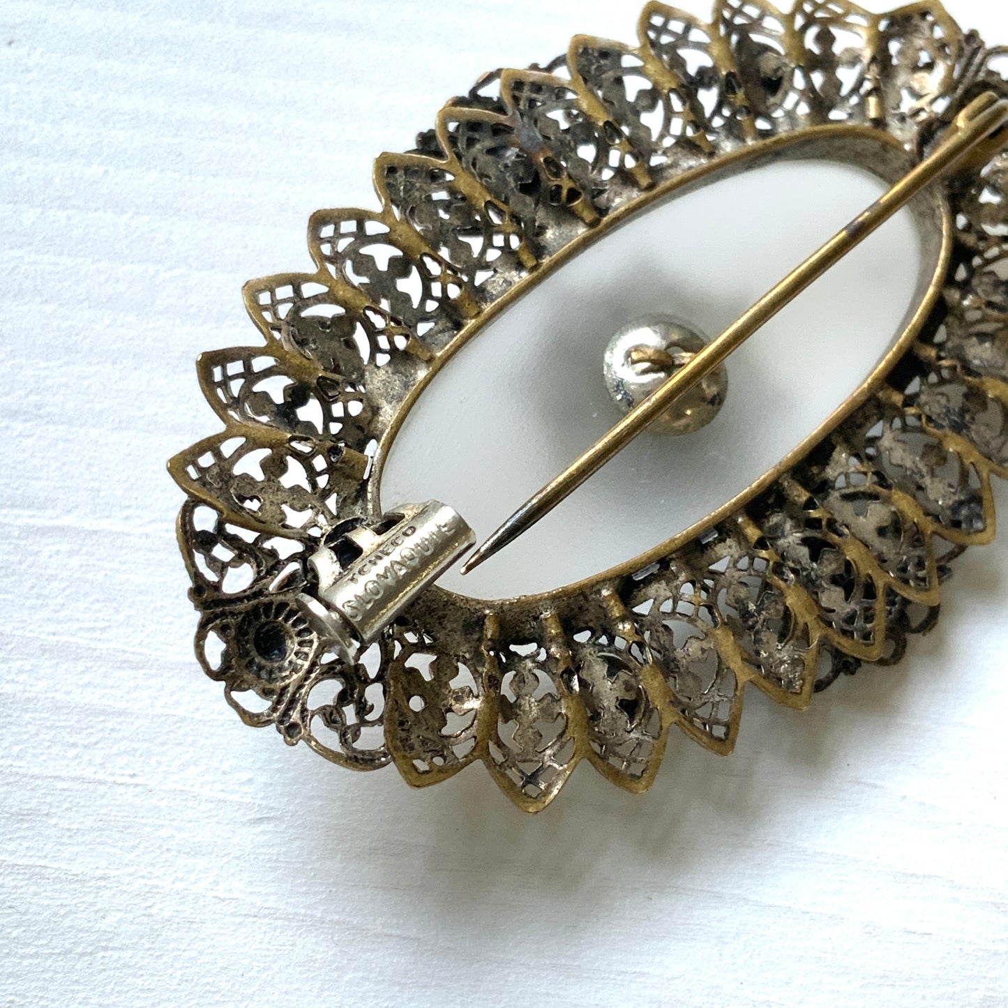 A brooch featuring a white oval frosted glass, framed with gold-tone filigree. 白いオーバルのフロスとグラスを、ゴールドトーンのフィリグリー縁取ったブローチ。
