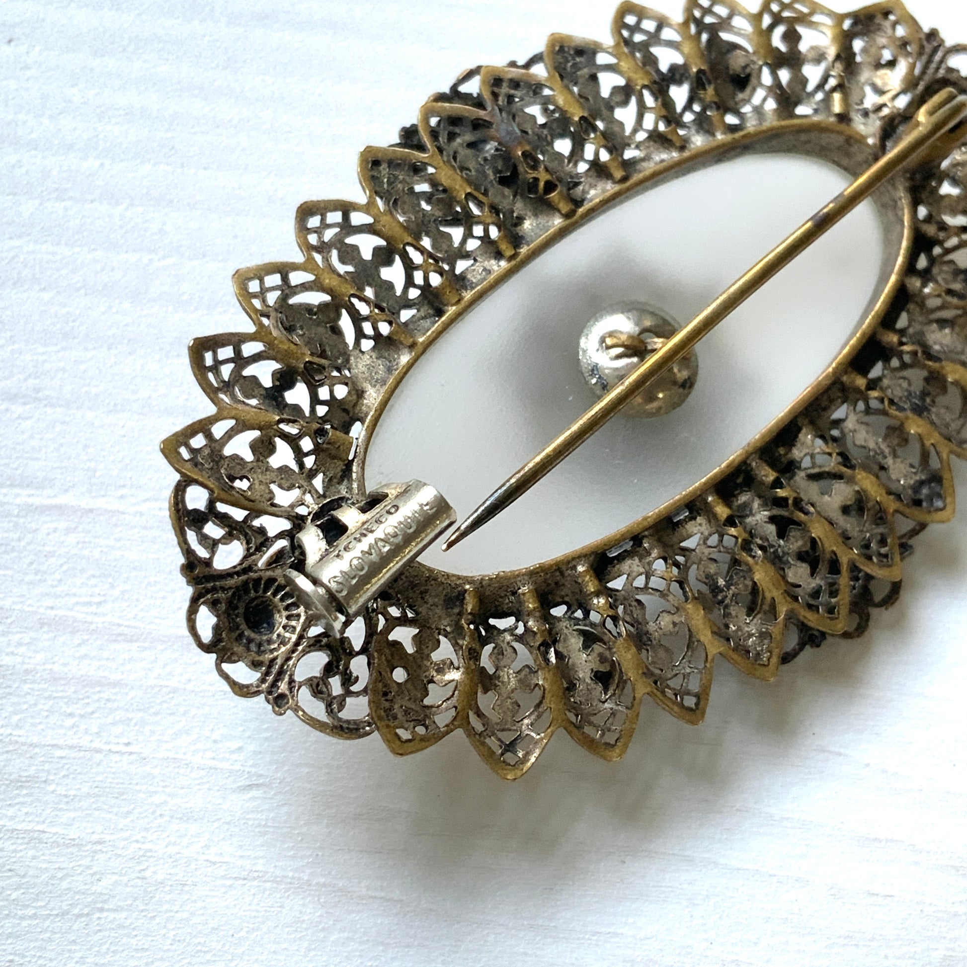 A brooch featuring a white oval frosted glass, framed with gold-tone filigree. 白いオーバルのフロスとグラスを、ゴールドトーンのフィリグリー縁取ったブローチ。