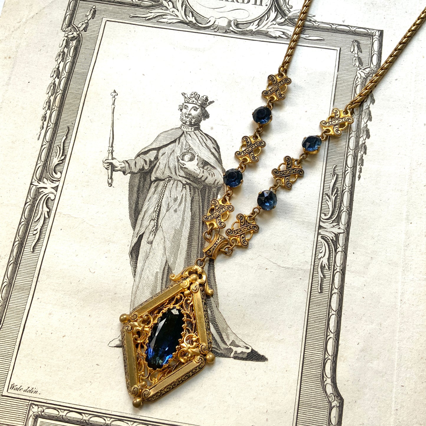 This necklace features a deep ocean-blue oval glass stone set at the center of a beautifully decorated diamond-shaped plate. 深い海のようなディープブルーのオーバルグラスが、美しい装飾を施したダイヤ型プレートの中央にあしらわれたネックレスです。