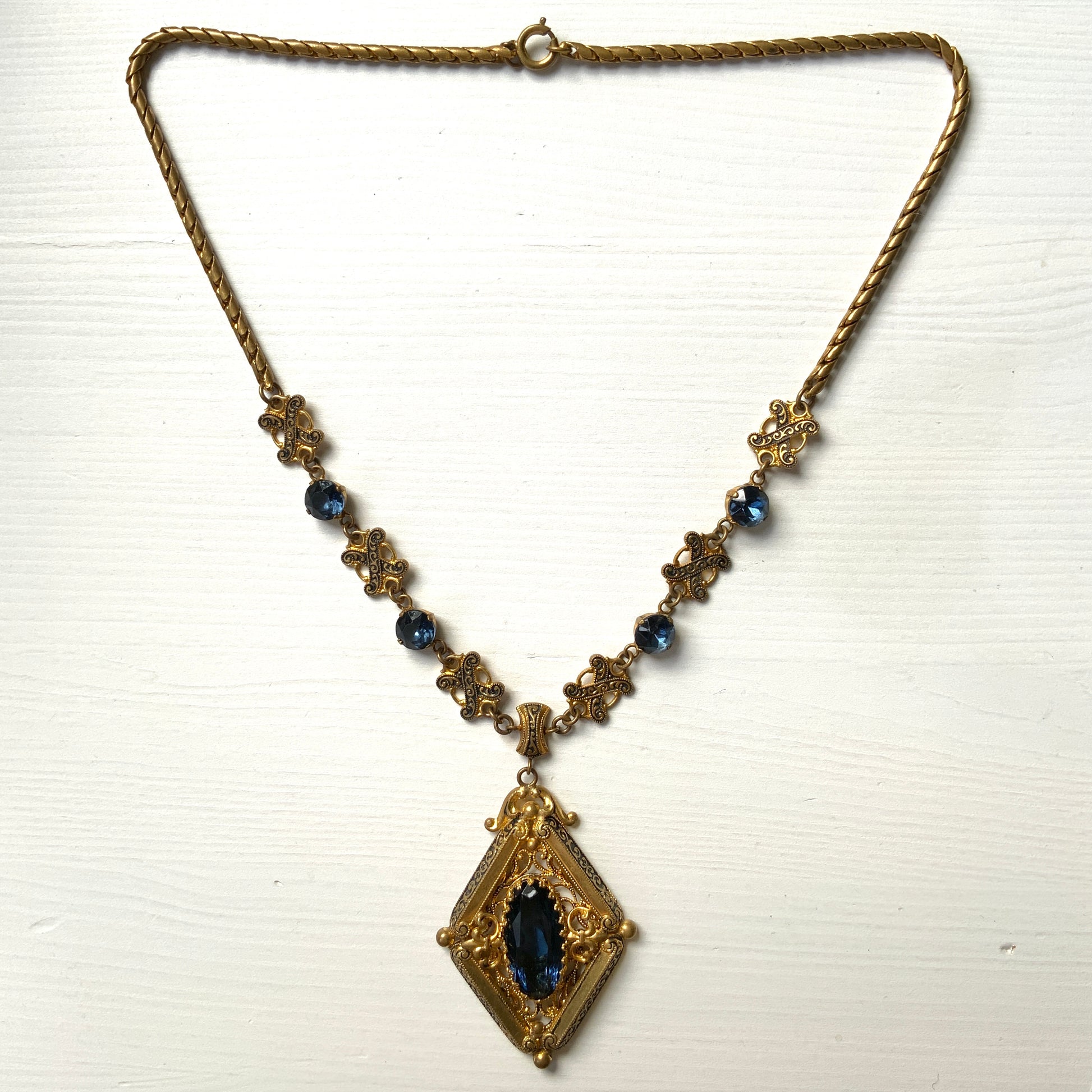This necklace features a deep ocean-blue oval glass stone set at the center of a beautifully decorated diamond-shaped plate. 深い海のようなディープブルーのオーバルグラスが、美しい装飾を施したダイヤ型プレートの中央にあしらわれたネックレスです。
