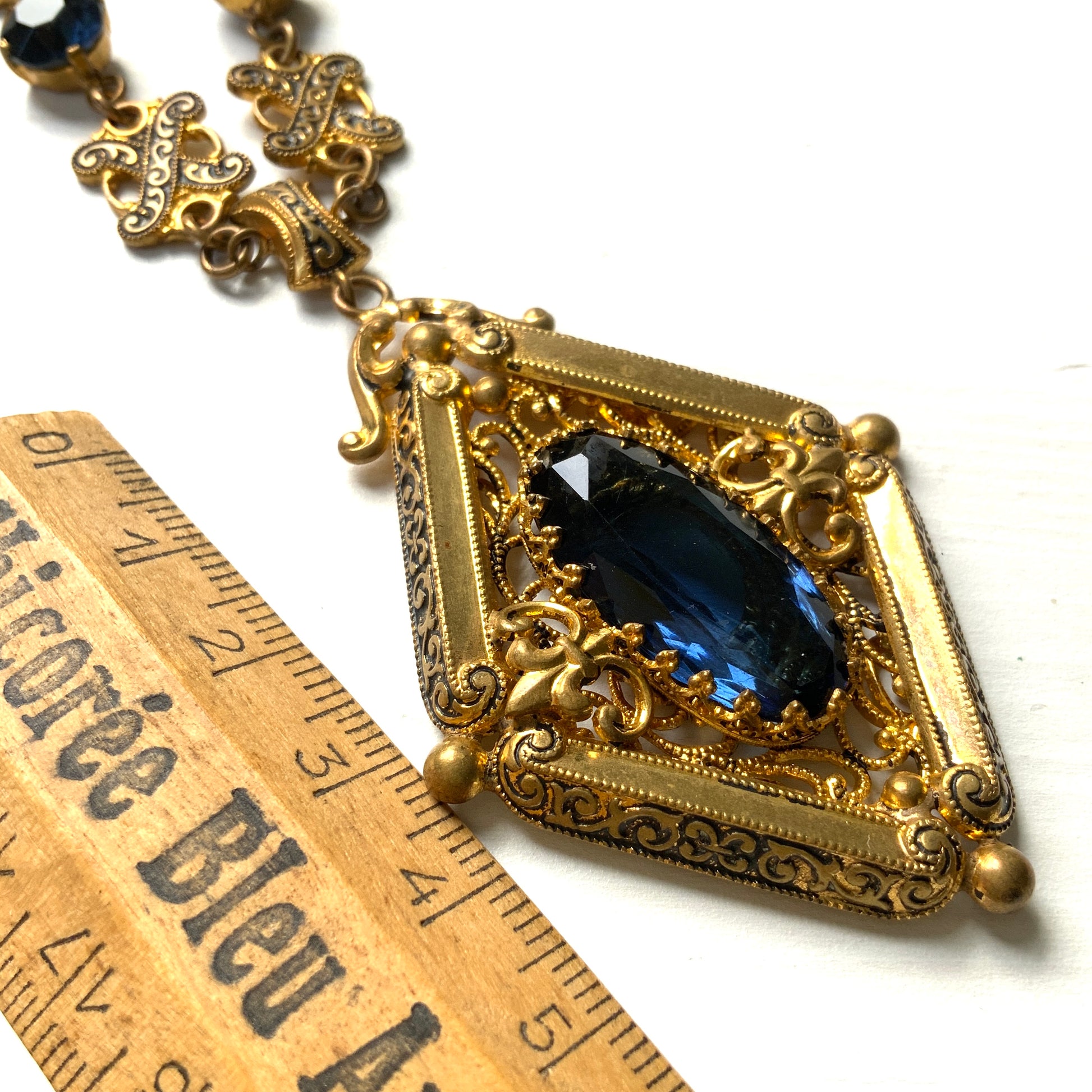 This necklace features a deep ocean-blue oval glass stone set at the center of a beautifully decorated diamond-shaped plate. 深い海のようなディープブルーのオーバルグラスが、美しい装飾を施したダイヤ型プレートの中央にあしらわれたネックレスです。