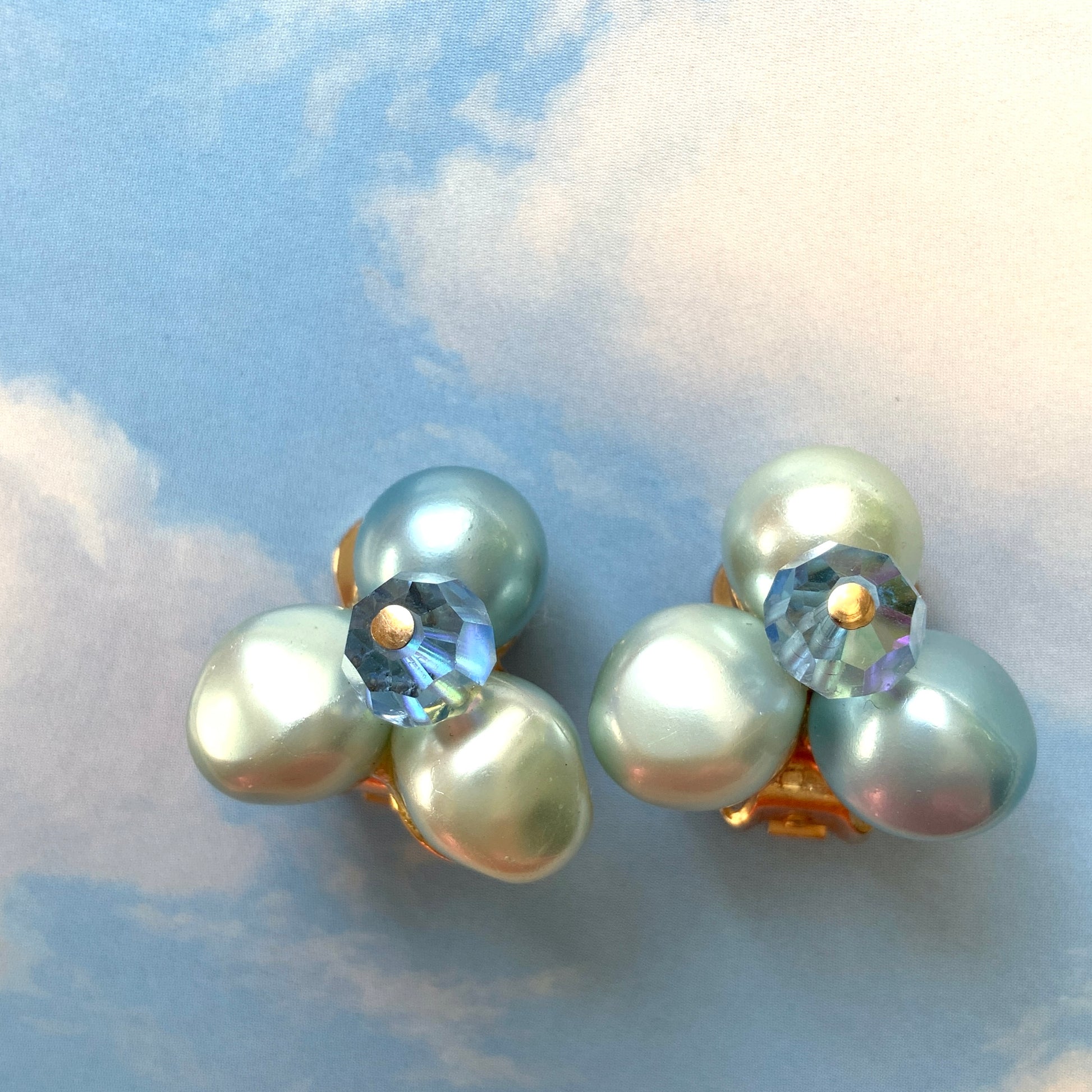 Clip-on earrings featuring three faux pearls in varying shades of blue, accented with a sparkling blue crystal bead at the center.3つの違うシェイドのブルーカラーパールがついたクリップイヤリング。