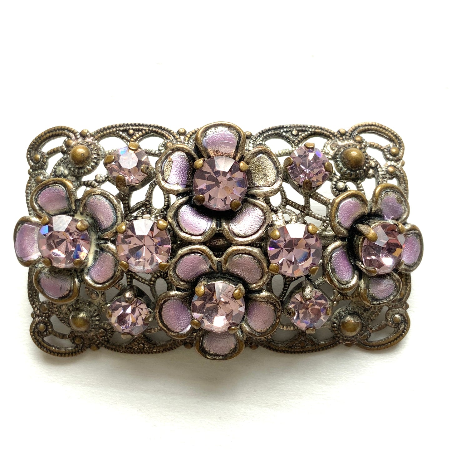 A brooch featuring a lilac-colorued flower and large matching rhinestones set on a rectangular base made of delicate filigree. 繊細なフィリグリーでできた長方形のベースに、ライラックカラーのお花と大粒の同色のラインストーンが光るブローチ。
