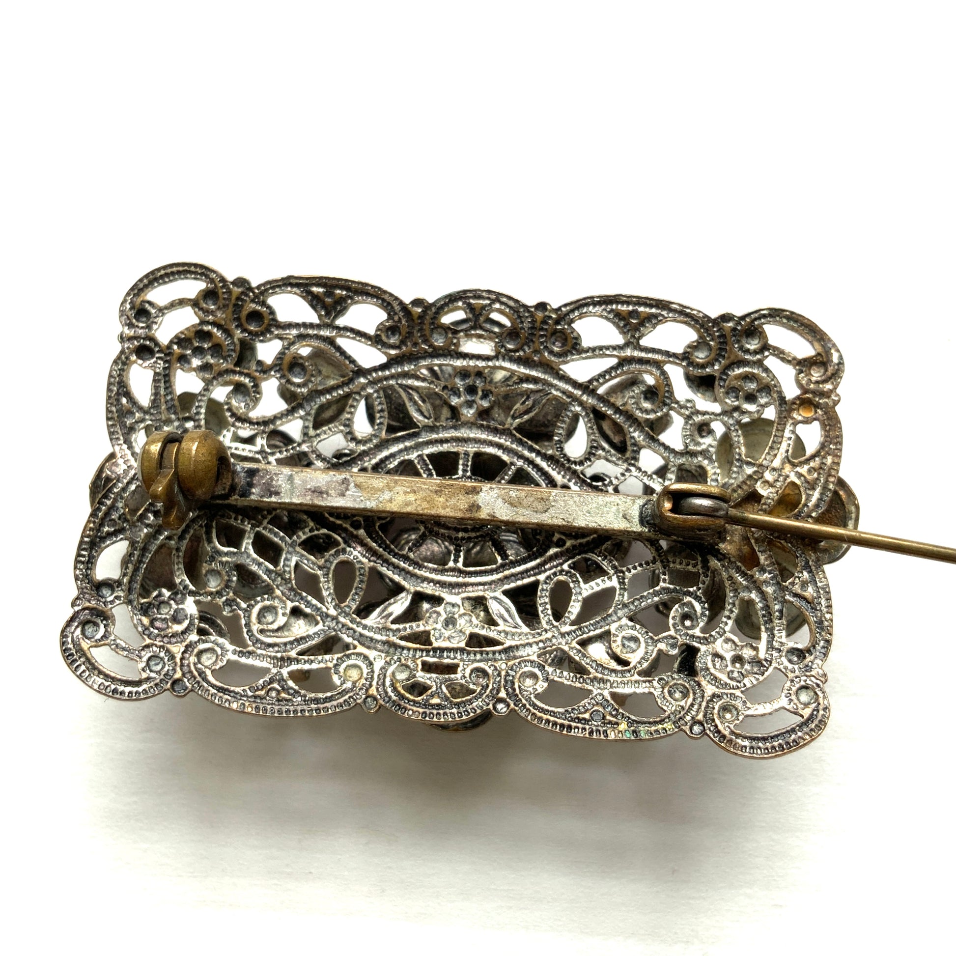 A brooch featuring a lilac-colorued flower and large matching rhinestones set on a rectangular base made of delicate filigree. 繊細なフィリグリーでできた長方形のベースに、ライラックカラーのお花と大粒の同色のラインストーンが光るブローチ。