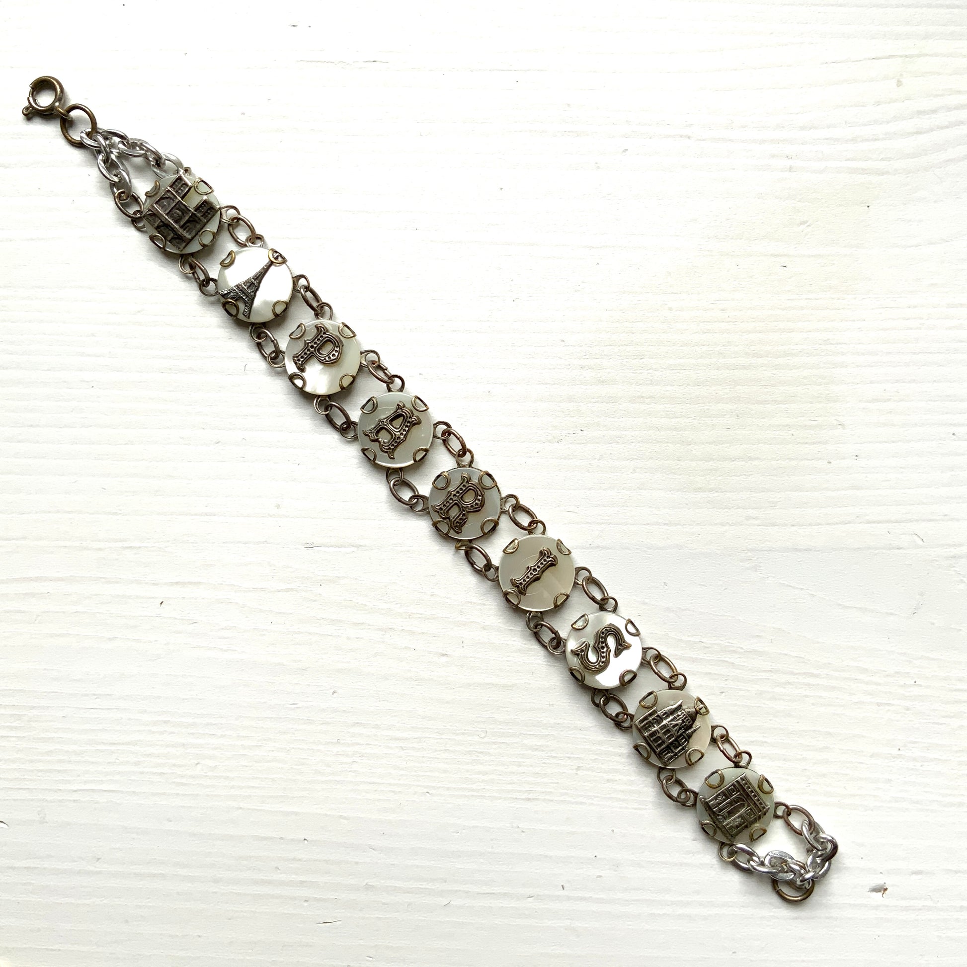 A bracelet featuring a mother of pearl tablet with the word "PARIS" and silver-tone charms of iconic Paris landmarks. マザーオブパールのタブレットに、PARISの文字とパリの観光名所のチャームがのったブレスレット。
