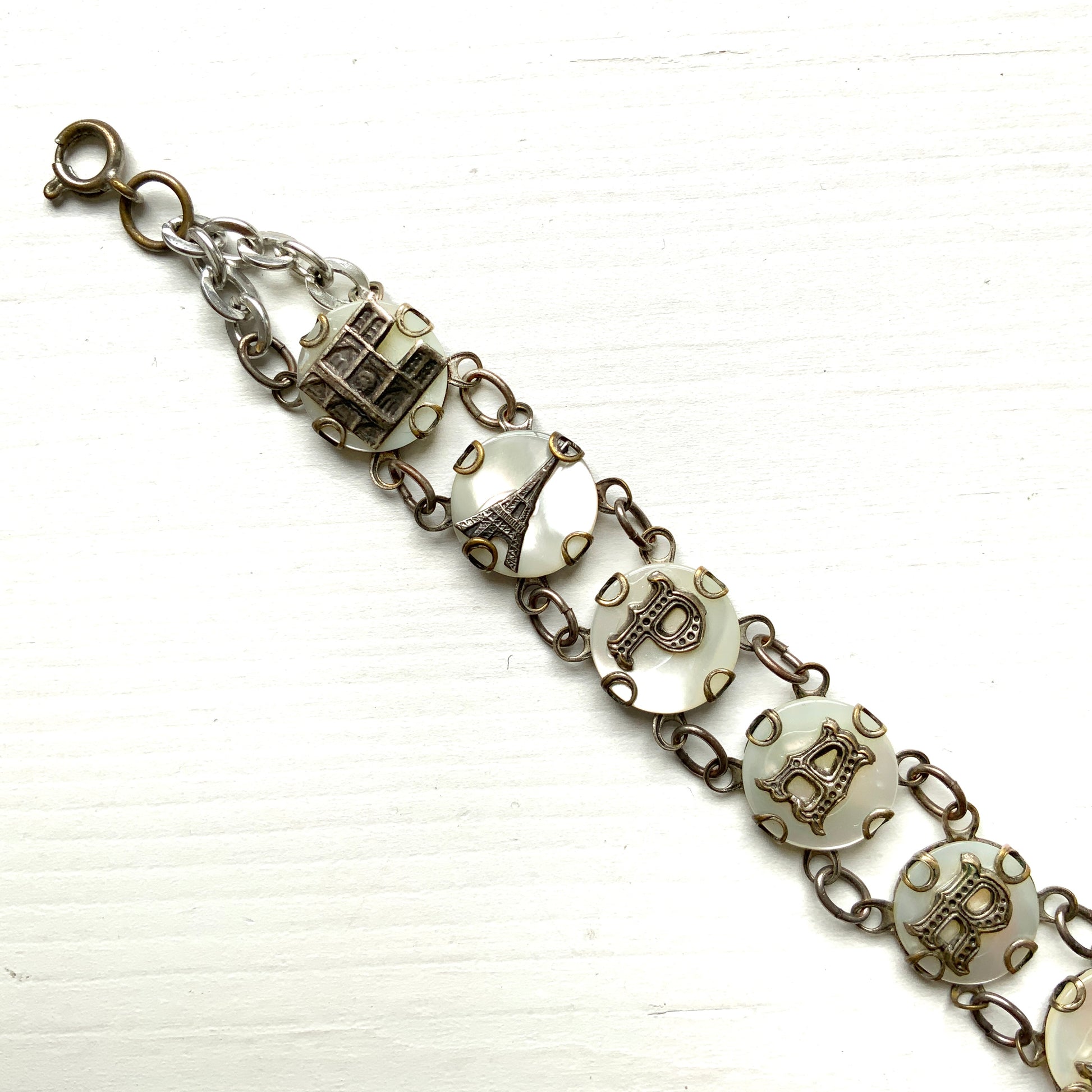 A bracelet featuring a mother of pearl tablet with the word "PARIS" and silver-tone charms of iconic Paris landmarks. マザーオブパールのタブレットに、PARISの文字とパリの観光名所のチャームがのったブレスレット。