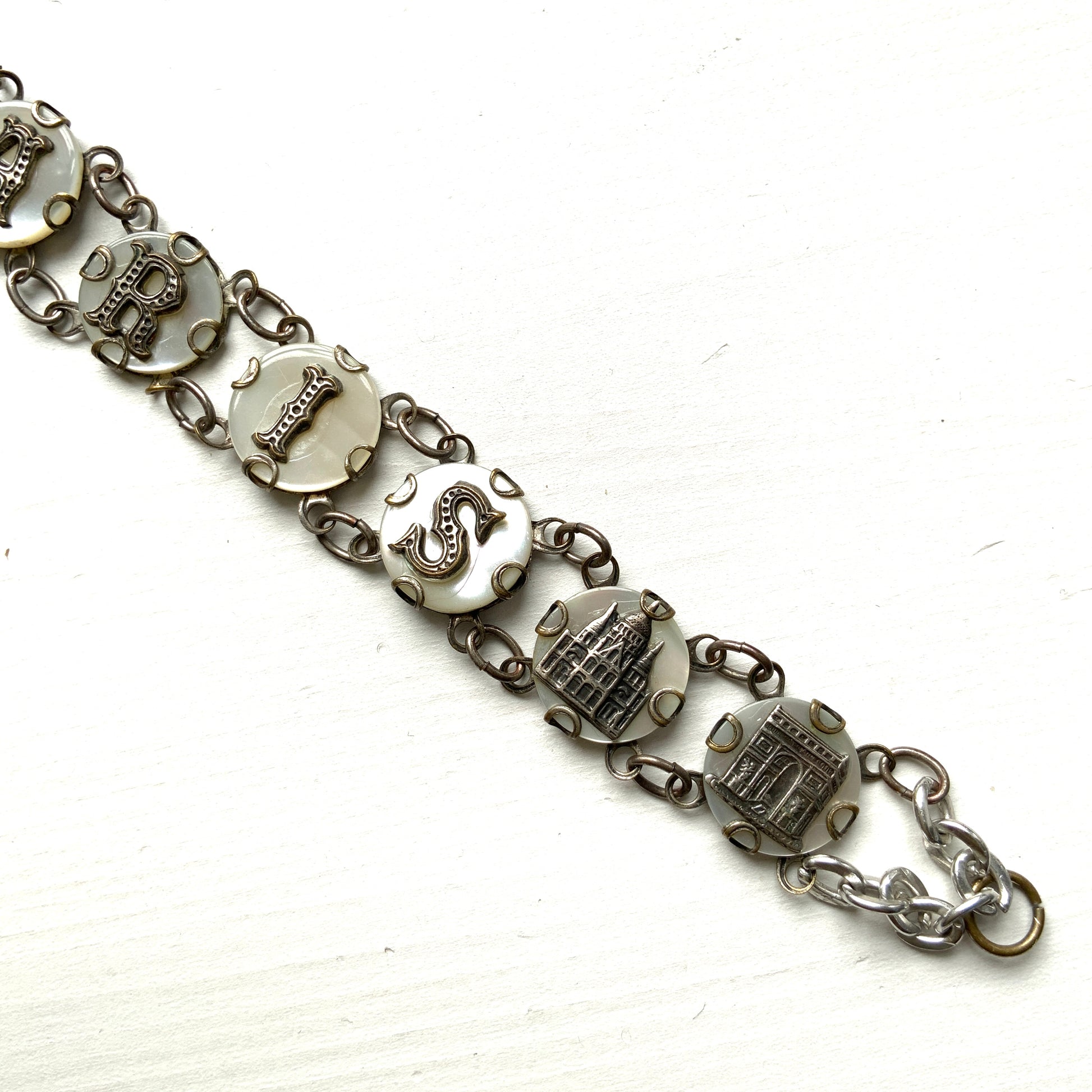 A bracelet featuring a mother of pearl tablet with the word "PARIS" and silver-tone charms of iconic Paris landmarks. マザーオブパールのタブレットに、PARISの文字とパリの観光名所のチャームがのったブレスレット。