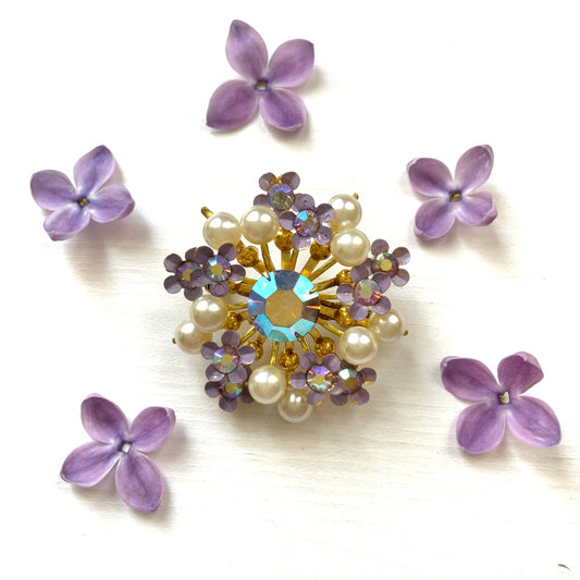 A brooch centered around large lilac-colored aurora borealis, adorned with small lilac enamel flowers and glass pearls. 大粒のライラックカラーのオーロラストーンを中心に、ライラックカラーのエナメルの小花とグラスパールがあしらわれたブローチ。