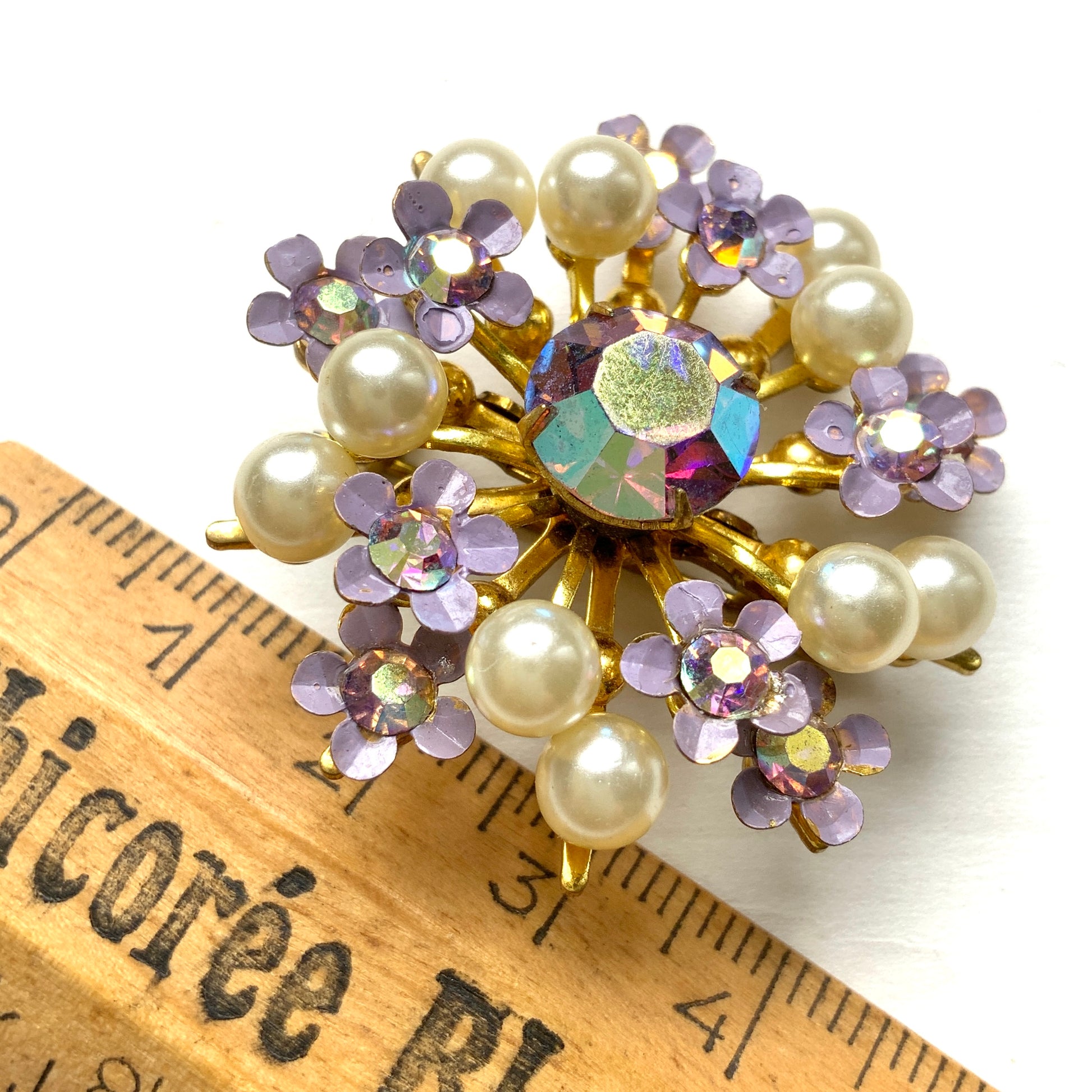 A brooch centered around large lilac-colored aurora borealis, adorned with small lilac enamel flowers and glass pearls. 大粒のライラックカラーのオーロラストーンを中心に、ライラックカラーのエナメルの小花とグラスパールがあしらわれたブローチ。