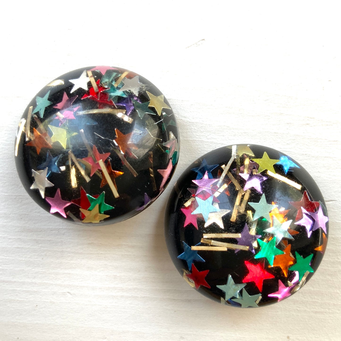 Clip-on earrings featuring a dome of black plastic filled with colourful star-shaped confetti. カラフルな星型コンフェッティが閉じ込められた、プラスチックドームのクリップイヤリング。