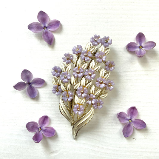 A brooch featuring charming light purple flowers resting on white enameled leaves. 白いエナメルの葉っぱにかわいらしい薄紫の小さなお花がたくさんついたブローチ。