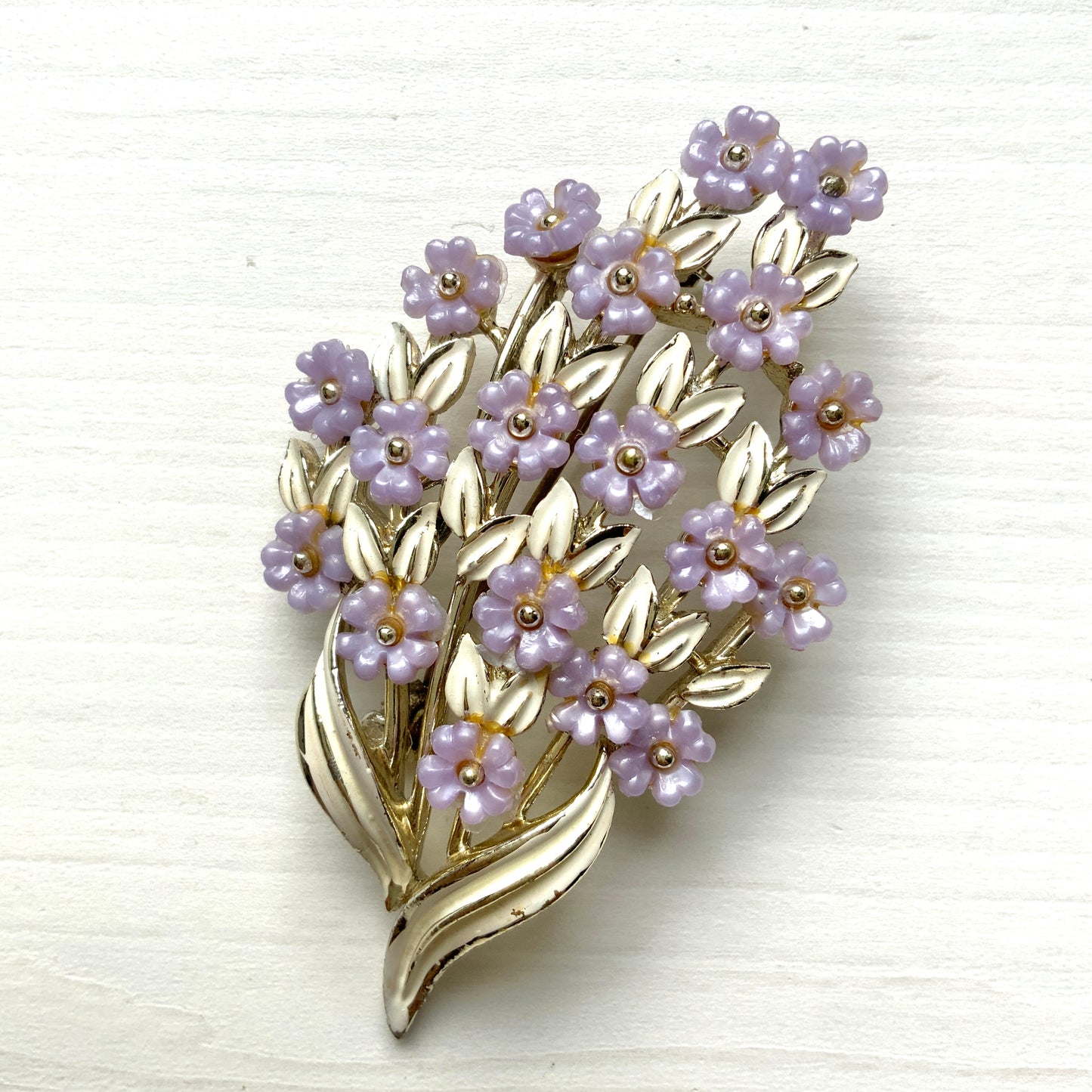 A brooch featuring charming light purple flowers resting on white enameled leaves. 白いエナメルの葉っぱにかわいらしい薄紫の小さなお花がたくさんついたブローチ。