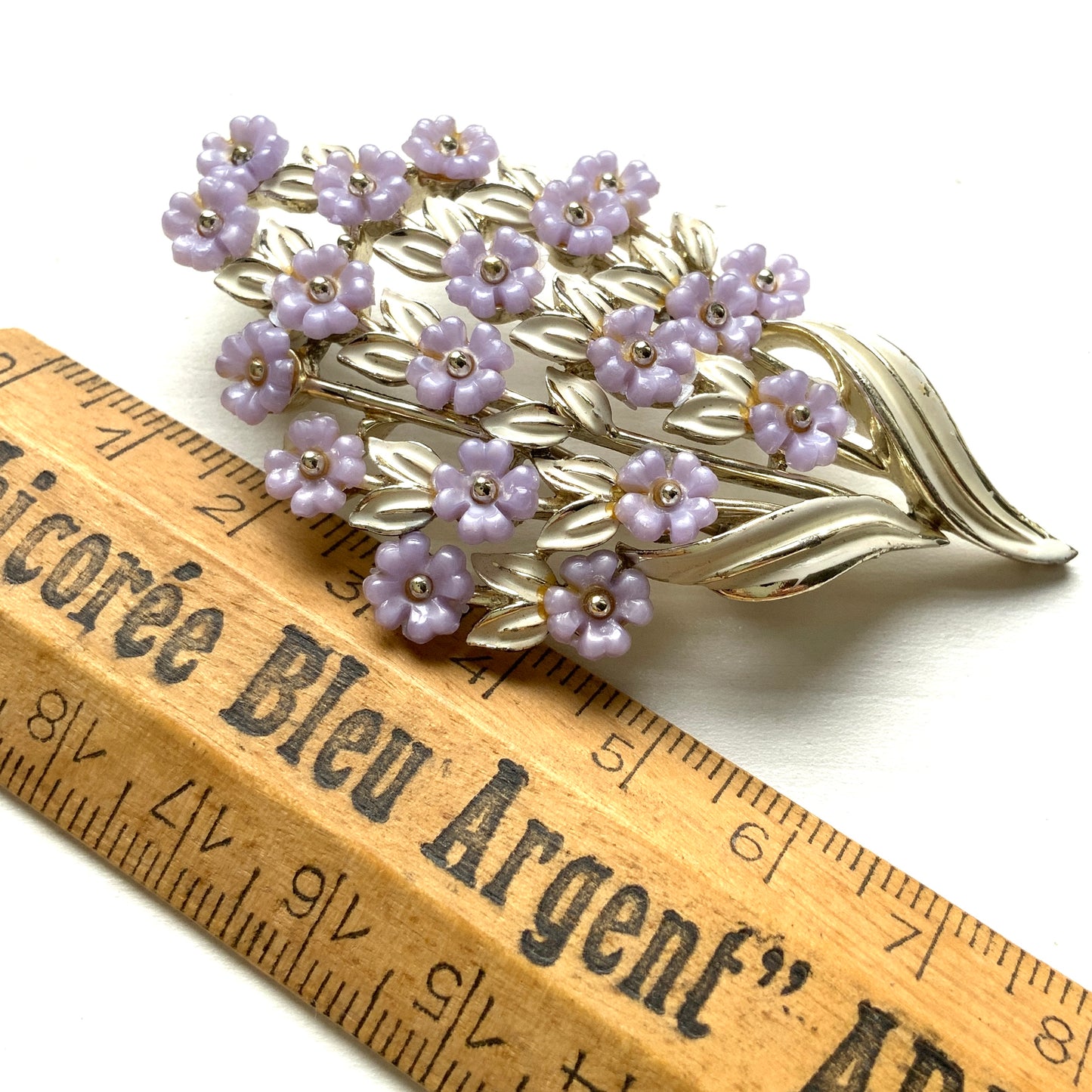 A brooch featuring charming light purple flowers resting on white enameled leaves. 白いエナメルの葉っぱにかわいらしい薄紫の小さなお花がたくさんついたブローチ。