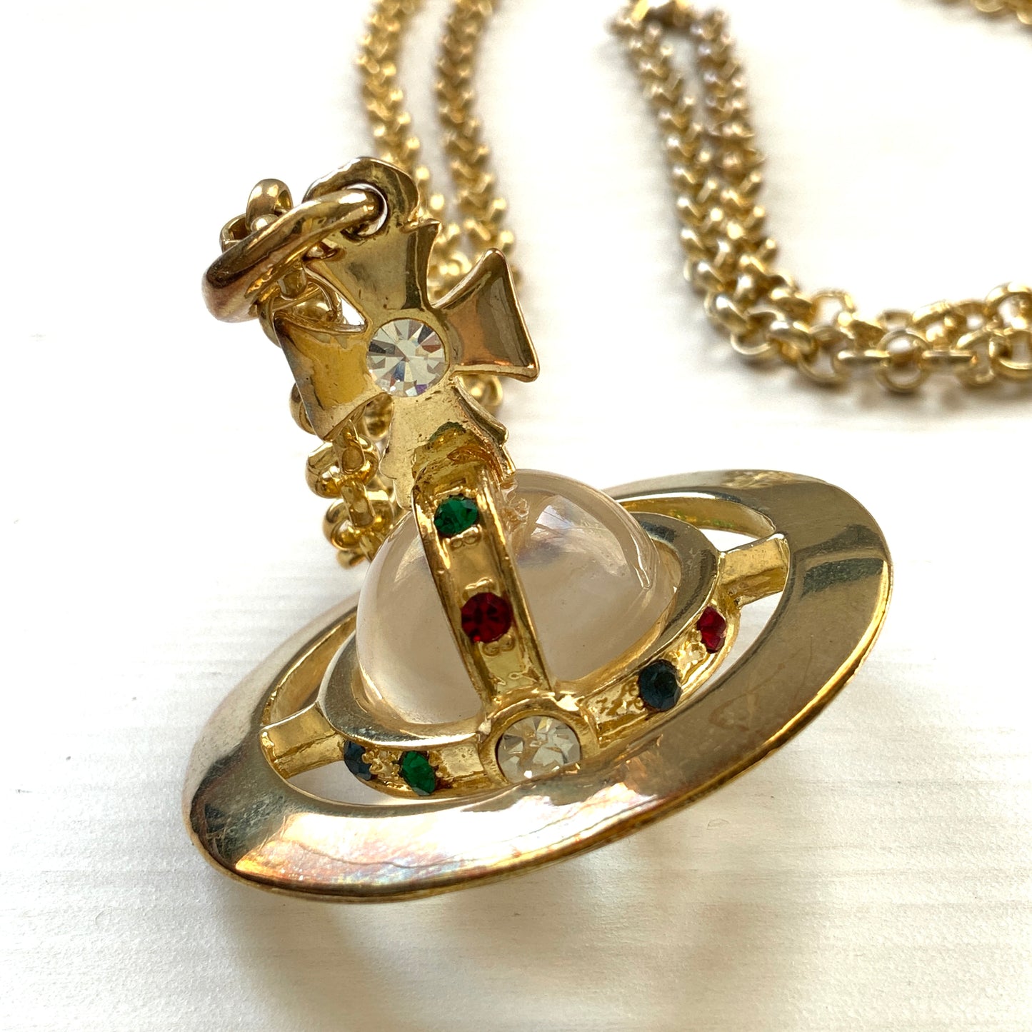 A striking pendant necklace featuring a decorative orb encircled by a metal hoop, resembling the rings of Saturn. カラフルなストーンで飾られた宝珠(キリスト教圏で、十字架をのせた球体として王権の象徴に用いられるモチーフ)を、まるで土星の輪のようなメタルフープで囲んだデザインのペンダントネックレスです。