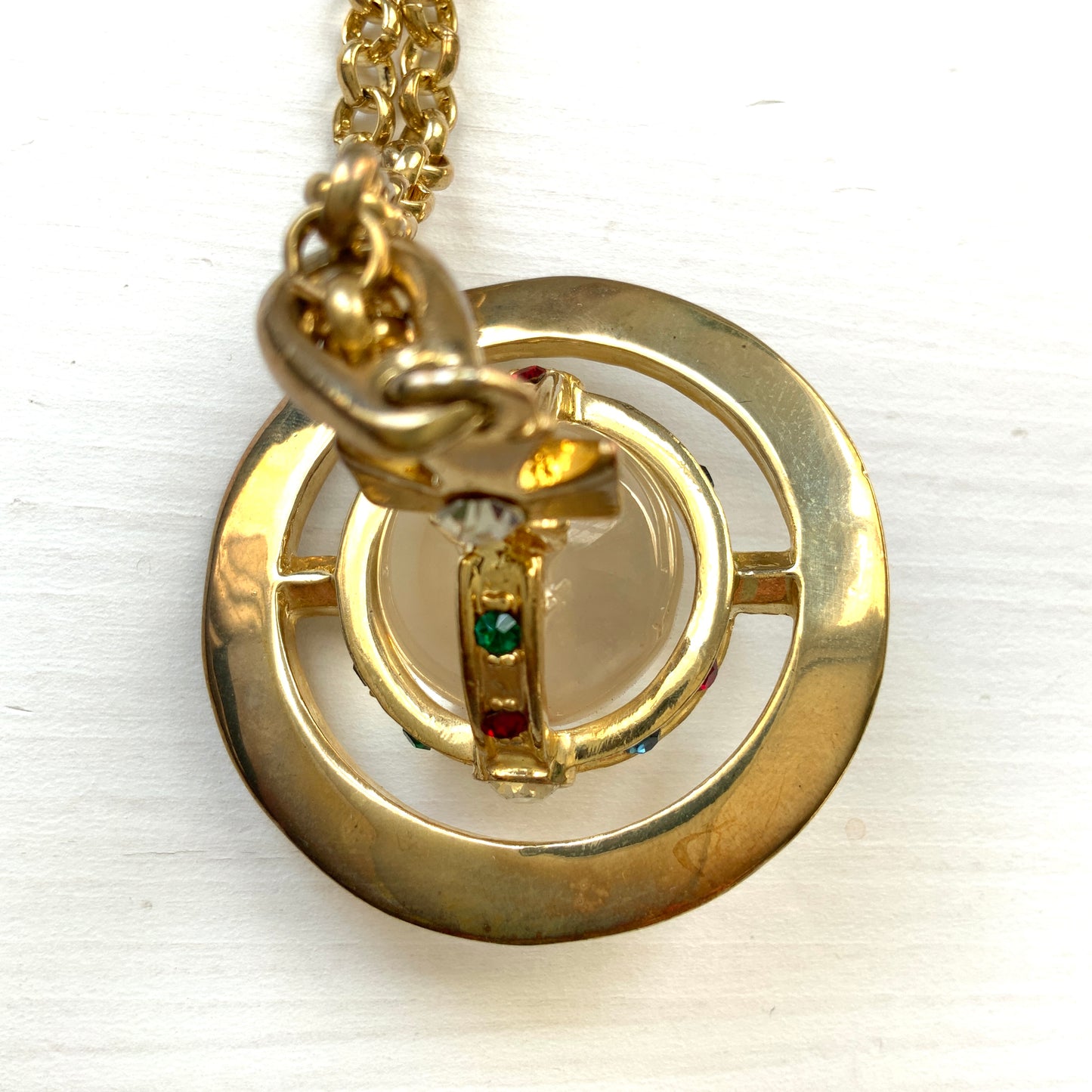 A striking pendant necklace featuring a decorative orb encircled by a metal hoop, resembling the rings of Saturn. カラフルなストーンで飾られた宝珠(キリスト教圏で、十字架をのせた球体として王権の象徴に用いられるモチーフ)を、まるで土星の輪のようなメタルフープで囲んだデザインのペンダントネックレスです。