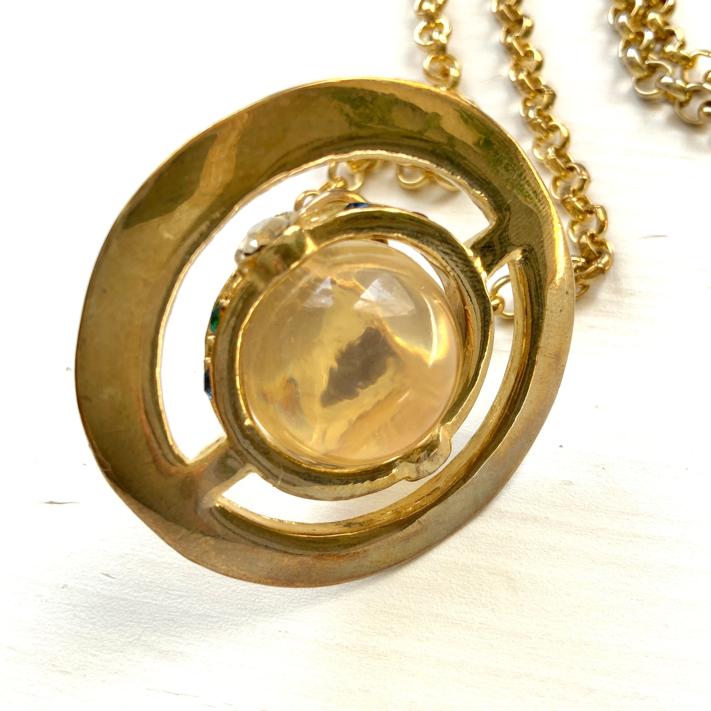 A striking pendant necklace featuring a decorative orb encircled by a metal hoop, resembling the rings of Saturn. カラフルなストーンで飾られた宝珠(キリスト教圏で、十字架をのせた球体として王権の象徴に用いられるモチーフ)を、まるで土星の輪のようなメタルフープで囲んだデザインのペンダントネックレスです。