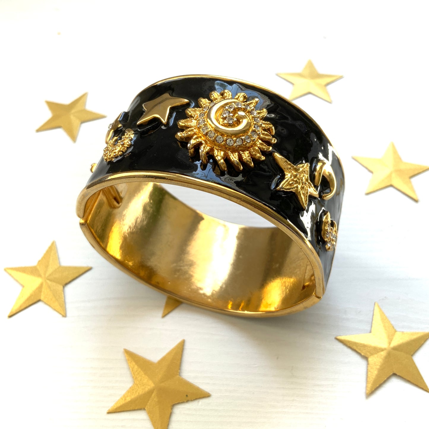 A wide bangle featuring celestial motifs in gold tone set against glossy black enamel. ブラックエナメルに、ゴールドトーンの天体モチーフが輝く幅広バングル。