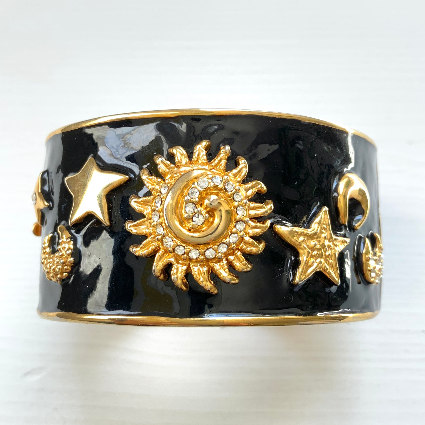 A wide bangle featuring celestial motifs in gold tone set against glossy black enamel. ブラックエナメルに、ゴールドトーンの天体モチーフが輝く幅広バングル。