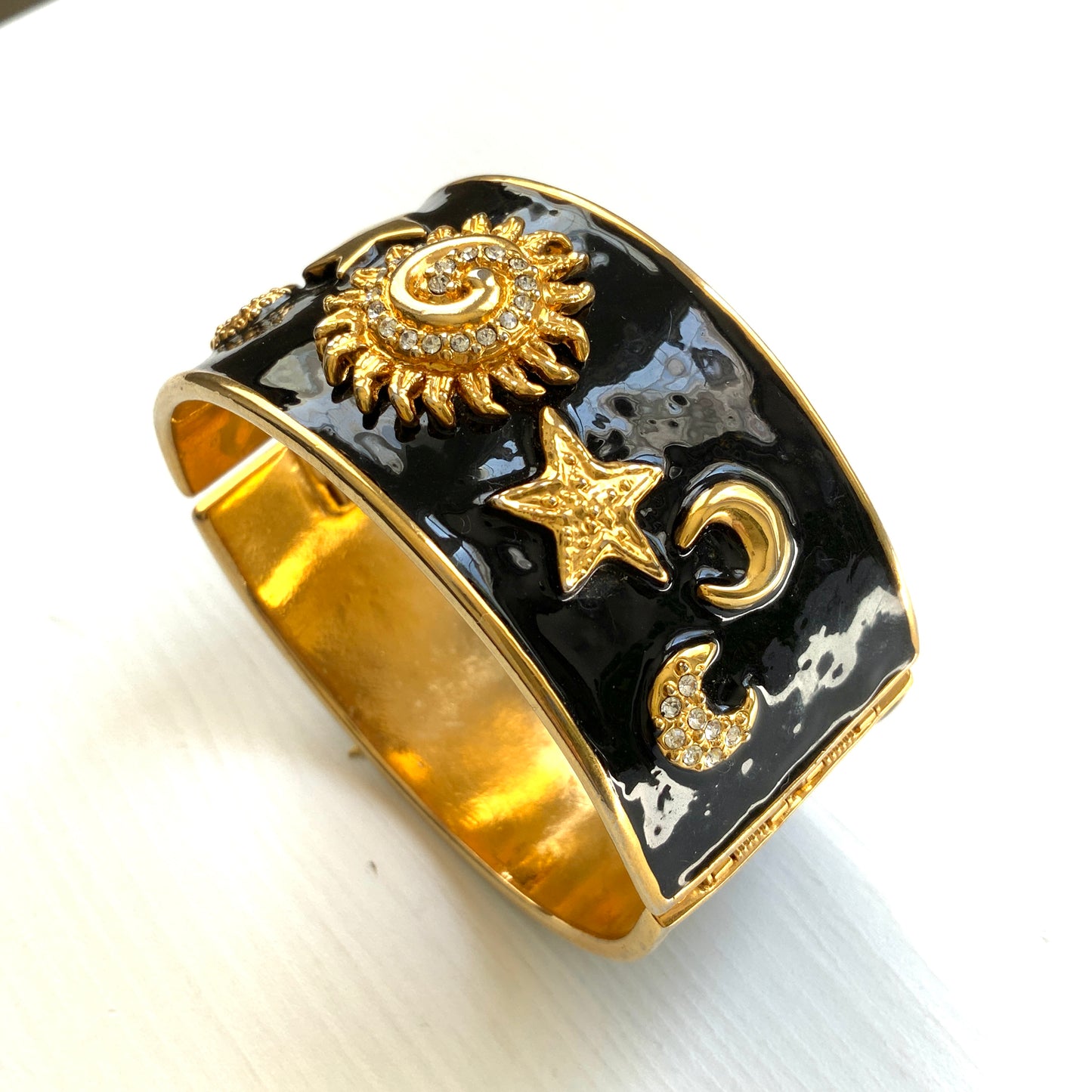 A wide bangle featuring celestial motifs in gold tone set against glossy black enamel. ブラックエナメルに、ゴールドトーンの天体モチーフが輝く幅広バングル。