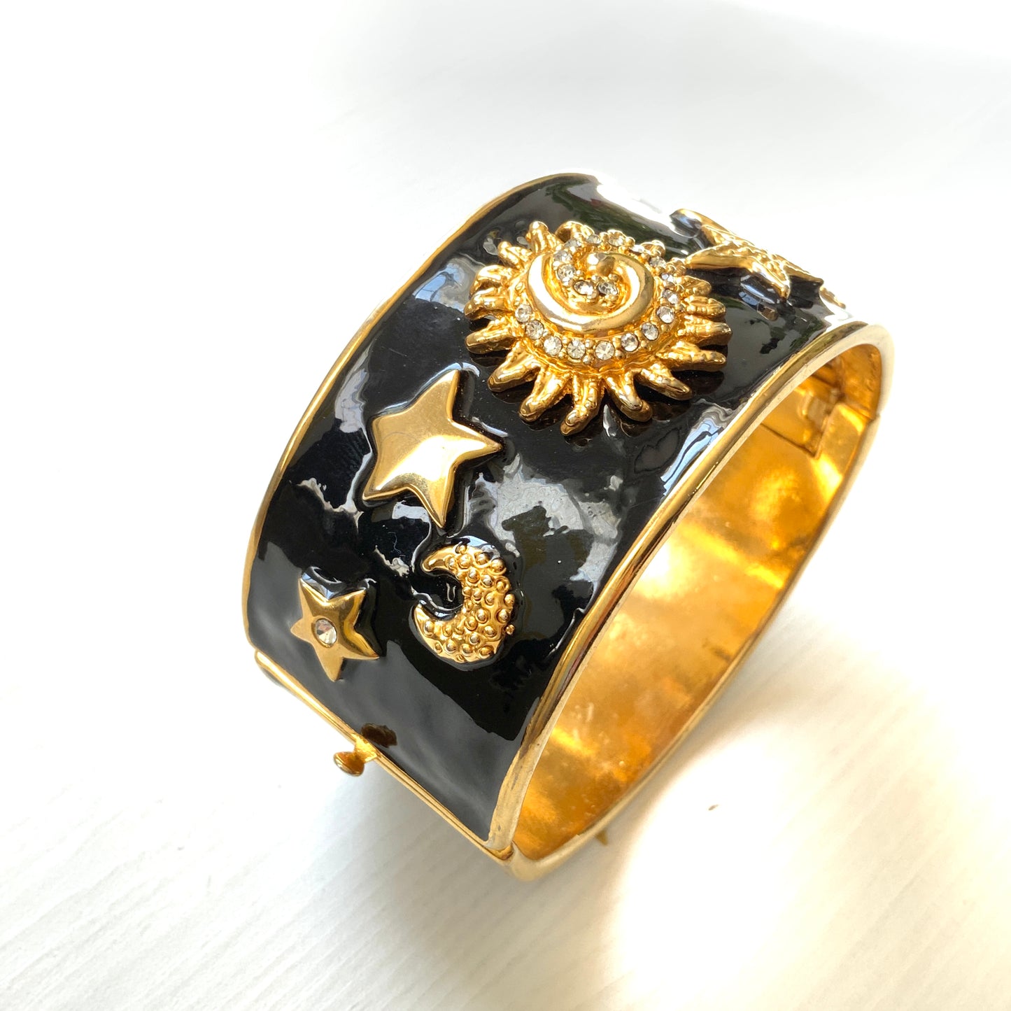 A wide bangle featuring celestial motifs in gold tone set against glossy black enamel. ブラックエナメルに、ゴールドトーンの天体モチーフが輝く幅広バングル。