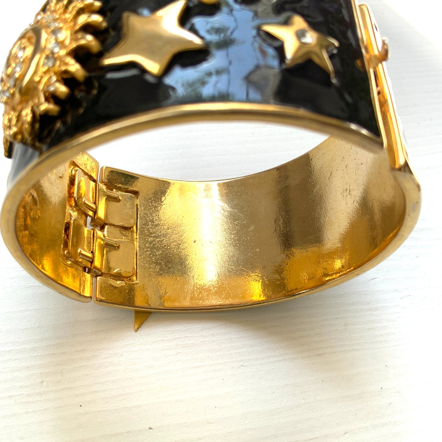 A wide bangle featuring celestial motifs in gold tone set against glossy black enamel. ブラックエナメルに、ゴールドトーンの天体モチーフが輝く幅広バングル。