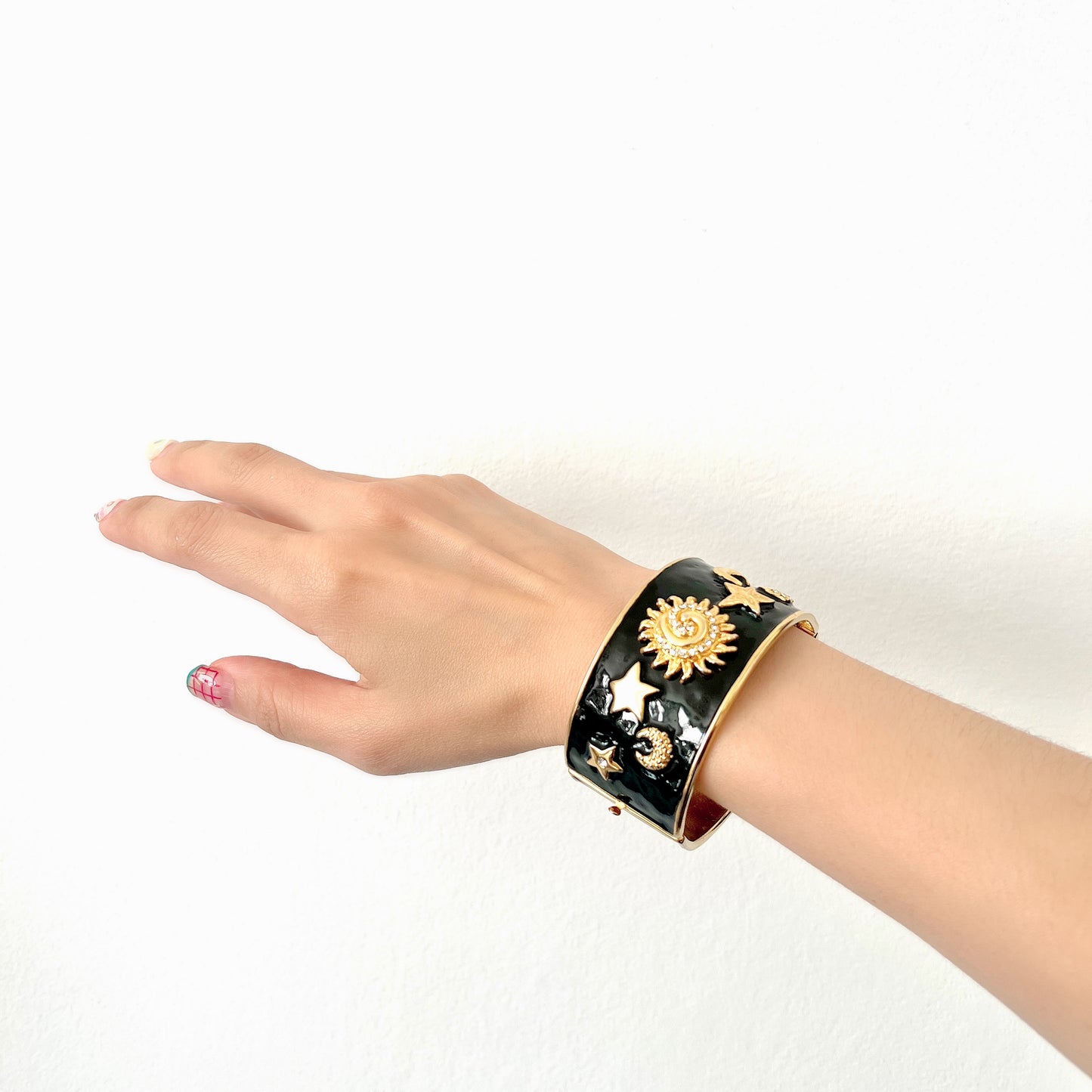 A wide bangle featuring celestial motifs in gold tone set against glossy black enamel. ブラックエナメルに、ゴールドトーンの天体モチーフが輝く幅広バングル。