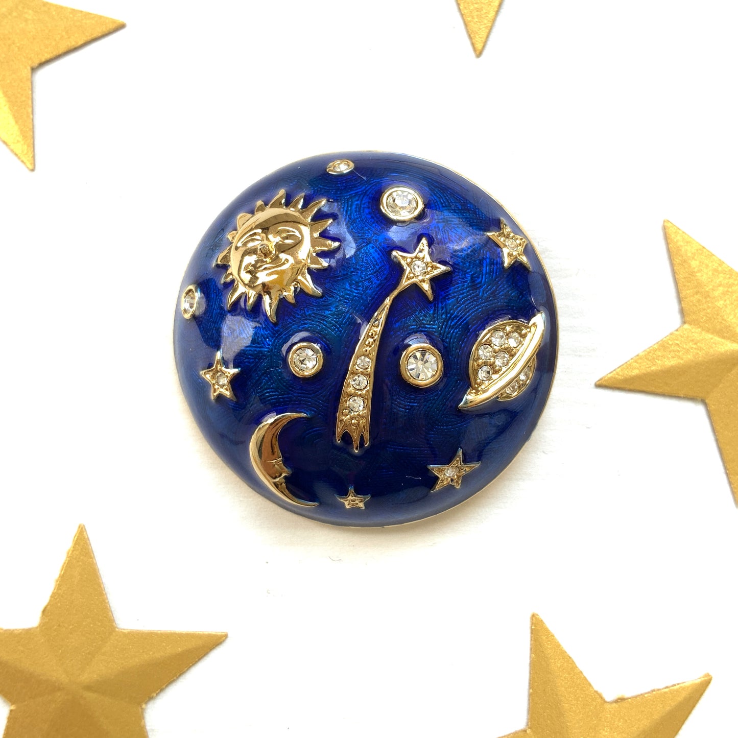 A dome-shaped brooch adorned with vibrant blue enamel, featuring silver-tone celestial motifs such as the sun face, crescent moon face, Saturn, and stars. 鮮やかなブルーのエナメルが施されたドームに、シルバートーンの太陽や三日月フェイスや、土星や星などが浮かんだブローチ。