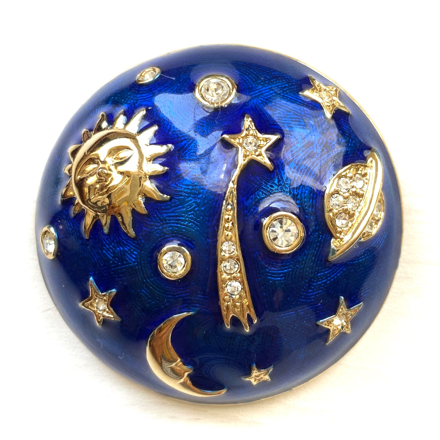 A dome-shaped brooch adorned with vibrant blue enamel, featuring silver-tone celestial motifs such as the sun face, crescent moon face, Saturn, and stars. 鮮やかなブルーのエナメルが施されたドームに、シルバートーンの太陽や三日月フェイスや、土星や星などが浮かんだブローチ。