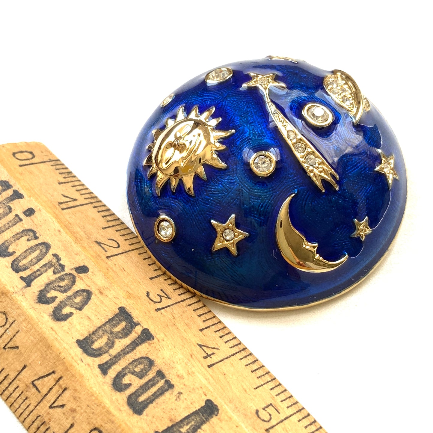 A dome-shaped brooch adorned with vibrant blue enamel, featuring silver-tone celestial motifs such as the sun face, crescent moon face, Saturn, and stars. 鮮やかなブルーのエナメルが施されたドームに、シルバートーンの太陽や三日月フェイスや、土星や星などが浮かんだブローチ。