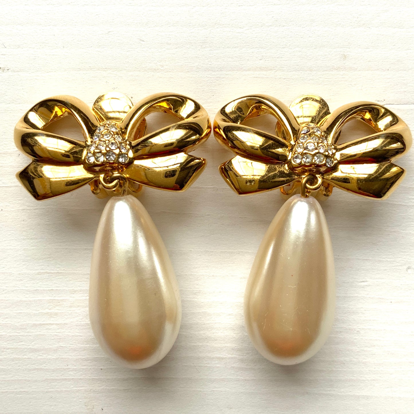 A pair of clip-on earrings featuring a bow motif in gold-tone metal, with large teardrop-shaped faux pearls that sway beautifully as you move. ゴールドトーンメタルのリボンモチーフに、大ぶりのティアドロップ型フォーパールが揺れるクリップイヤリング。