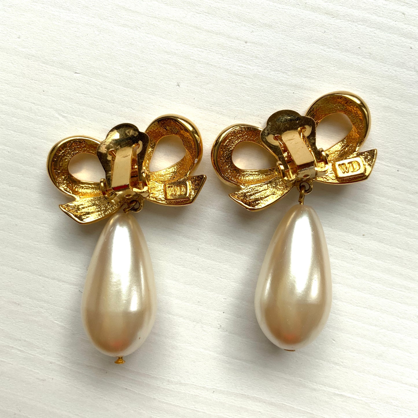 A pair of clip-on earrings featuring a bow motif in gold-tone metal, with large teardrop-shaped faux pearls that sway beautifully as you move. ゴールドトーンメタルのリボンモチーフに、大ぶりのティアドロップ型フォーパールが揺れるクリップイヤリング。