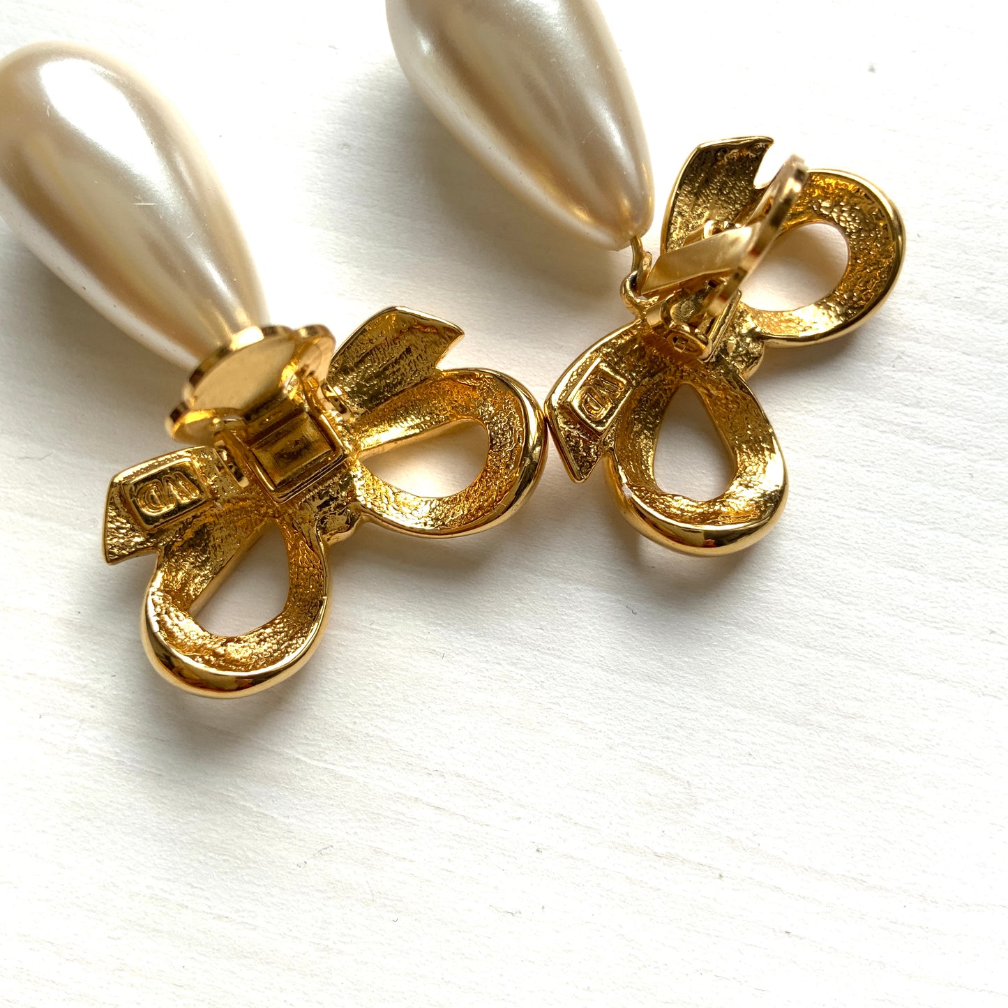 A pair of clip-on earrings featuring a bow motif in gold-tone metal, with large teardrop-shaped faux pearls that sway beautifully as you move. ゴールドトーンメタルのリボンモチーフに、大ぶりのティアドロップ型フォーパールが揺れるクリップイヤリング。