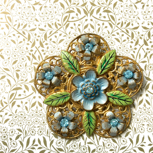 This brooch features a gold-tone floral filigree design, delicately painted with soft blue enamel flowers.  ゴールドトーンのお花の形のフィリグリーに、淡いブルーでエナメルペイントされたお花が描かれたブローチ。