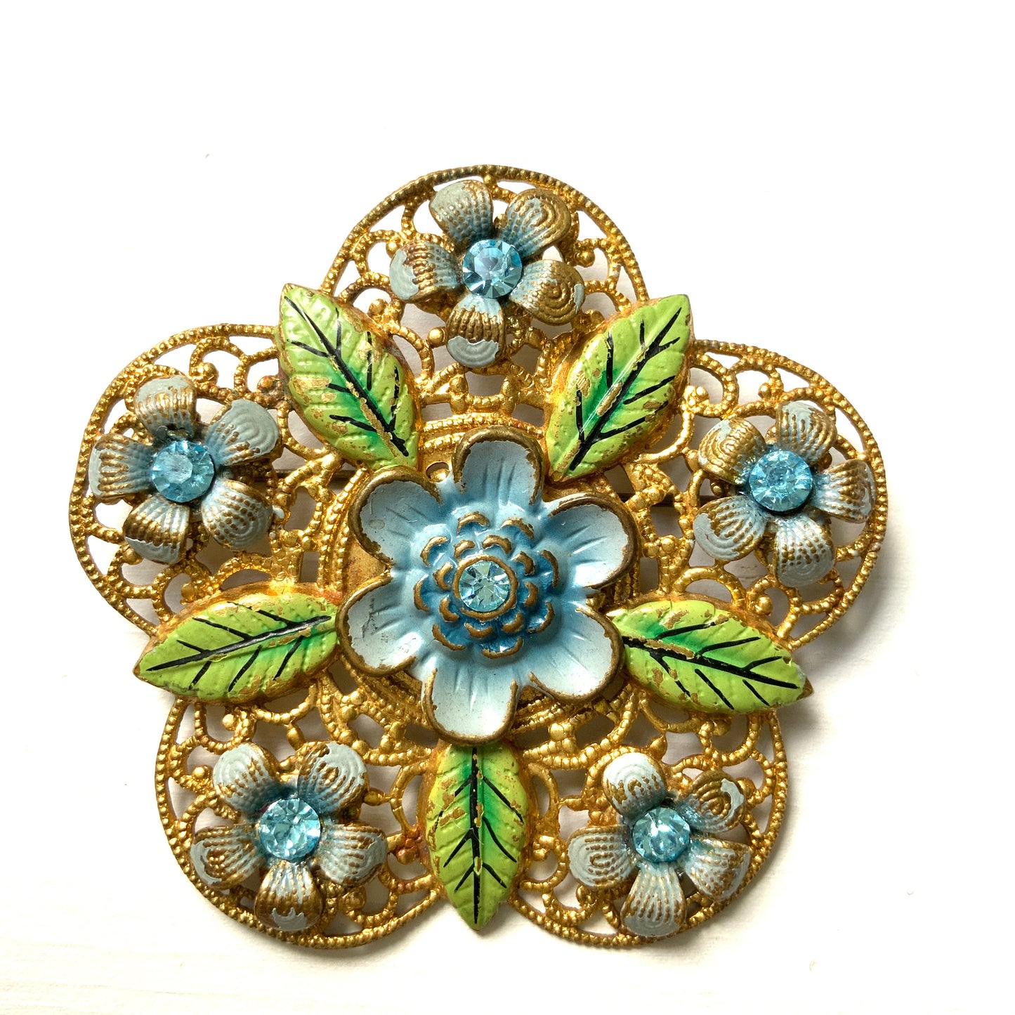 This brooch features a gold-tone floral filigree design, delicately painted with soft blue enamel flowers.  ゴールドトーンのお花の形のフィリグリーに、淡いブルーでエナメルペイントされたお花が描かれたブローチ。