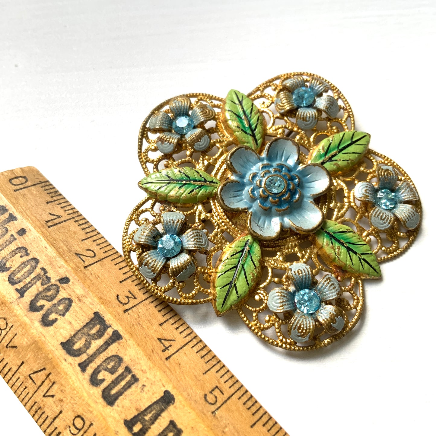 This brooch features a gold-tone floral filigree design, delicately painted with soft blue enamel flowers.  ゴールドトーンのお花の形のフィリグリーに、淡いブルーでエナメルペイントされたお花が描かれたブローチ。