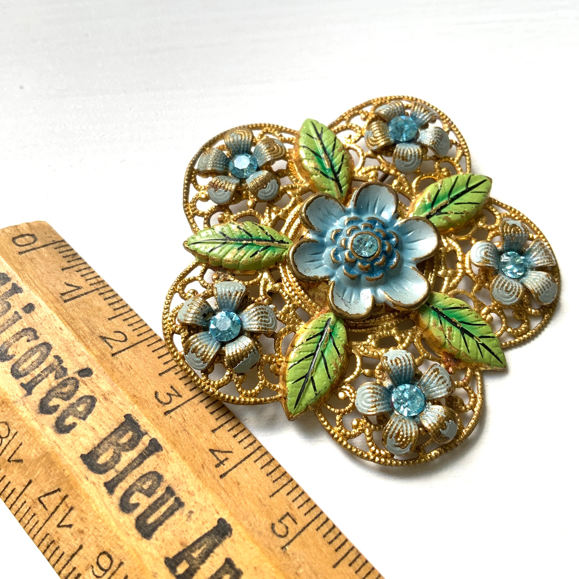 This brooch features a gold-tone floral filigree design, delicately painted with soft blue enamel flowers.  ゴールドトーンのお花の形のフィリグリーに、淡いブルーでエナメルペイントされたお花が描かれたブローチ。