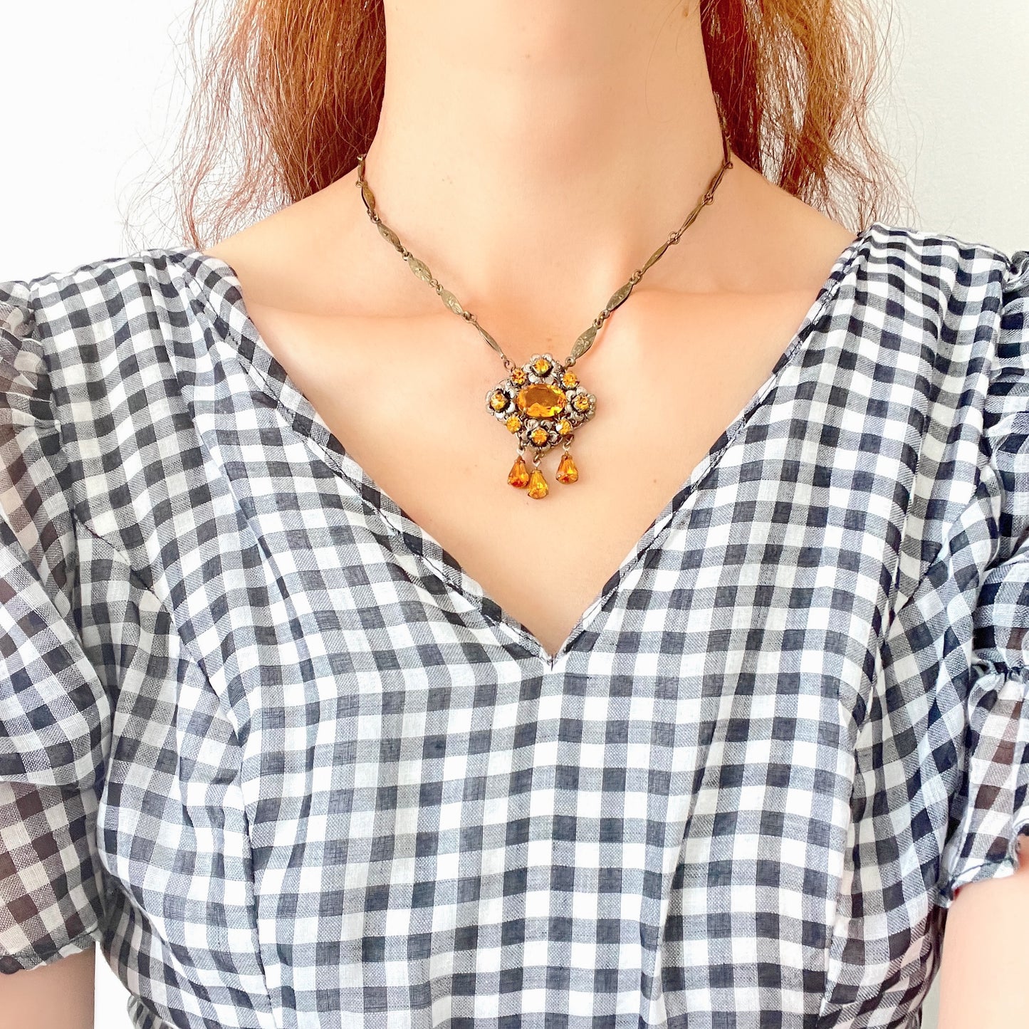 An oval amber glass jewel is surrounded by delicate metal flowers, with matching amber teardrop rhinestones gently dangling below. オーバル型のアンバーグラスビジューの周りをメタルのお花が取り囲み、同じアンバーカラーのティアドロップラインストーンが揺れるネックレス。